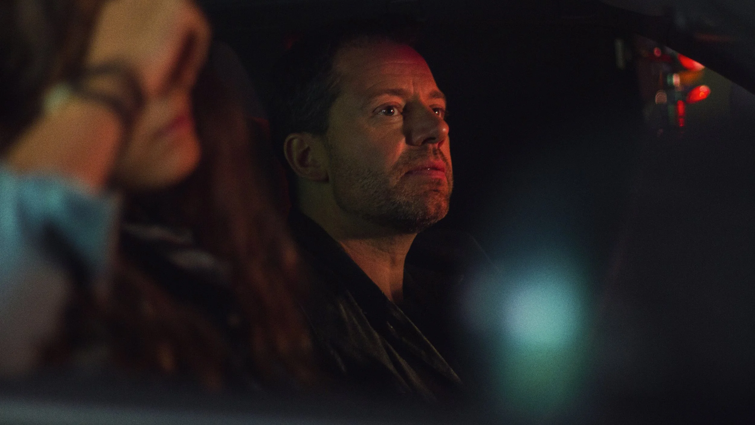 Un homme regarde sérieusement par une fenêtre dans un environnement sombre, avec un éclairage tamisé.