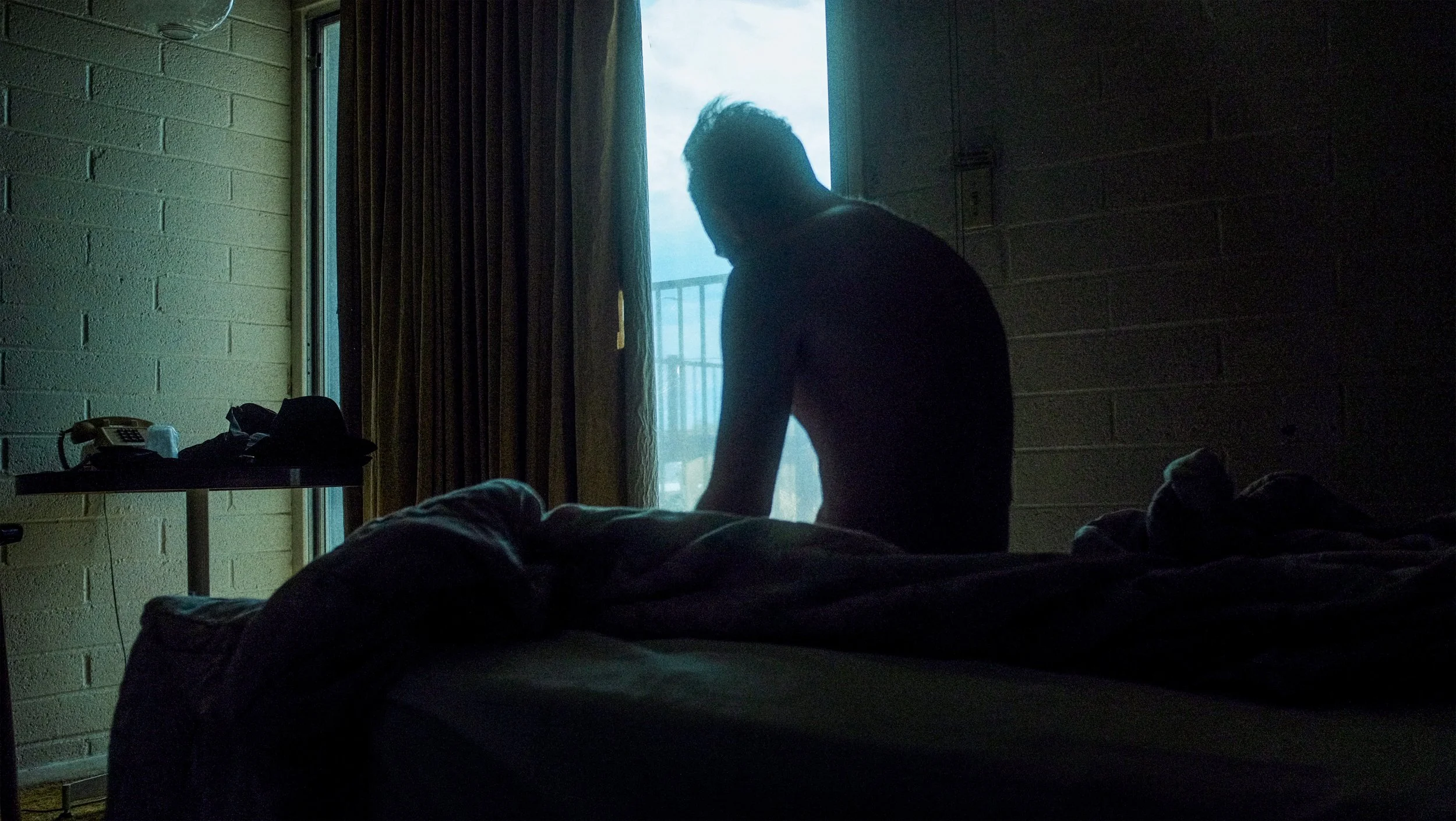 Silhouette d'une personne assise au bord du lit dans une chambre sombre, avec une lumière provenant d'une porte ou d'une fenêtre ouverte à l'extérieur.