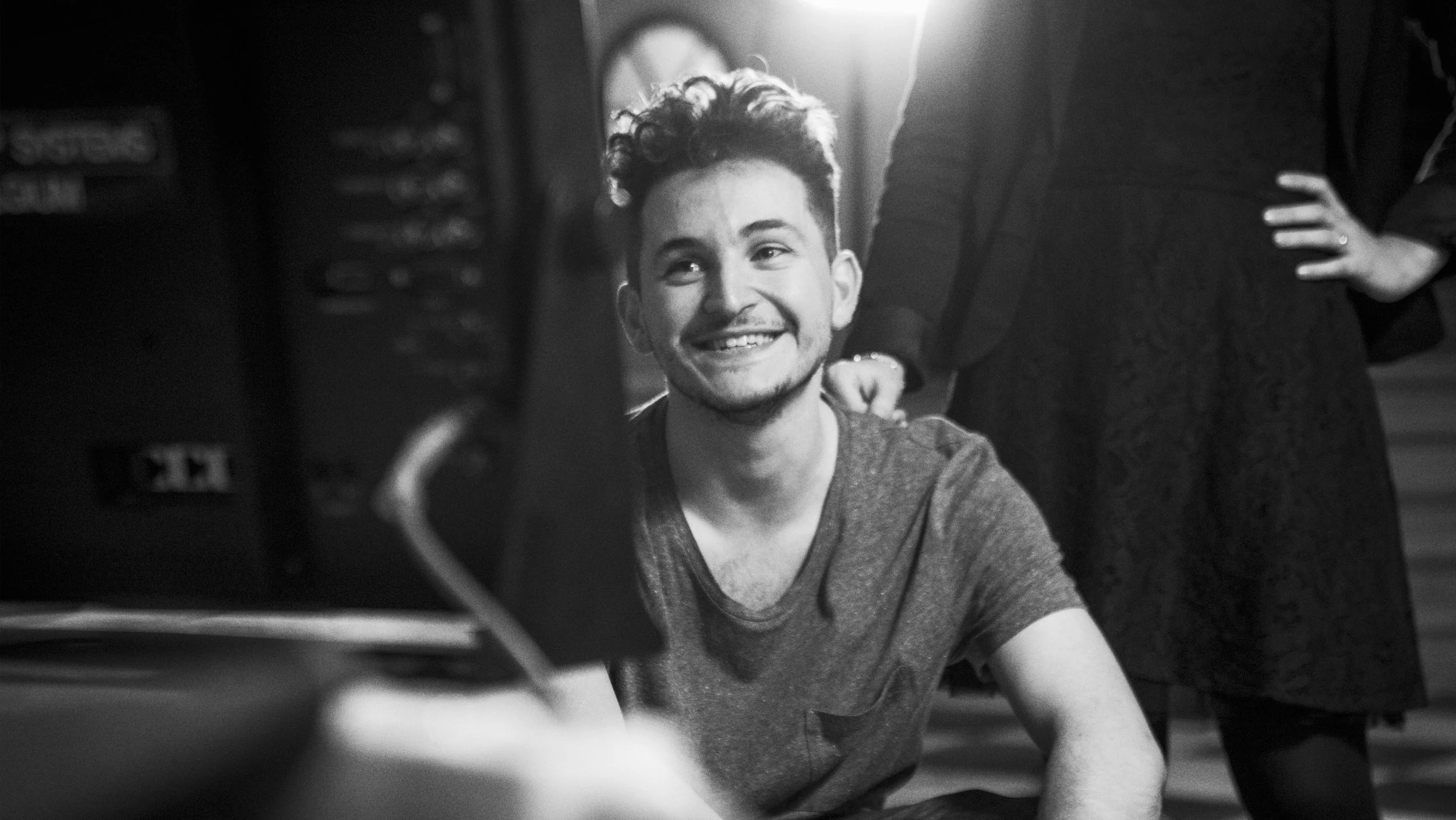 Un jeune homme souriant, assis, avec une personne debout à côté, dans un environnement intérieur.