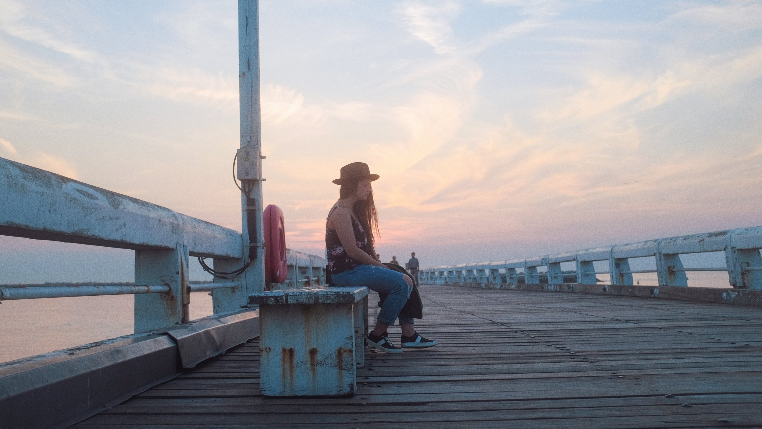 Femme assise sur un banc en bois, portant un chapeau noir, regardant vers le bas, sur une jetée en bois au coucher du soleil, avec un ciel coloré.