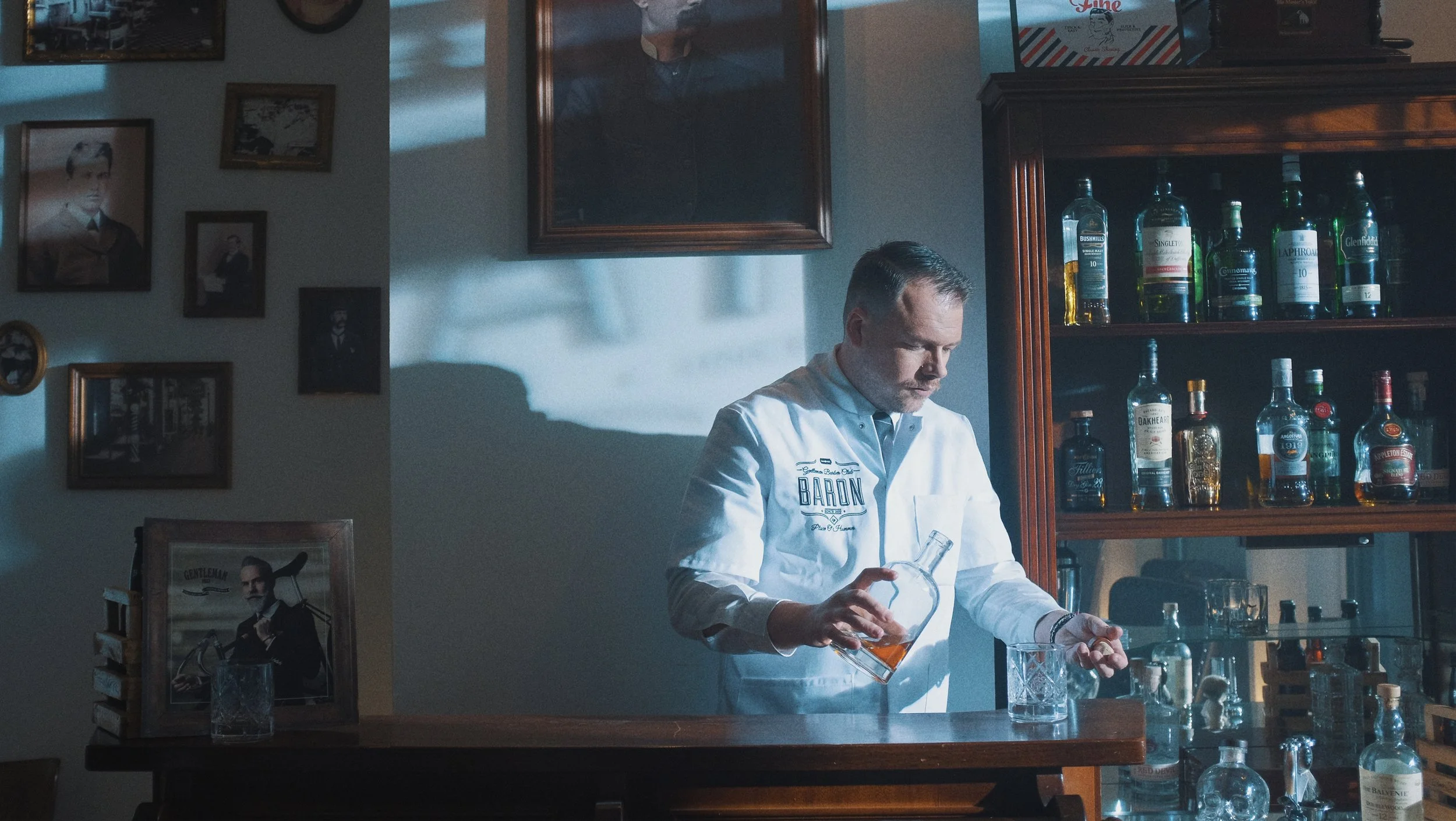 Un homme en chemise blanche versant un whisky dans un verre dans un bar, avec des bouteilles d'alcool dans une étagère en bois derrière lui, dans une pièce avec des portraits anciens sur le mur.