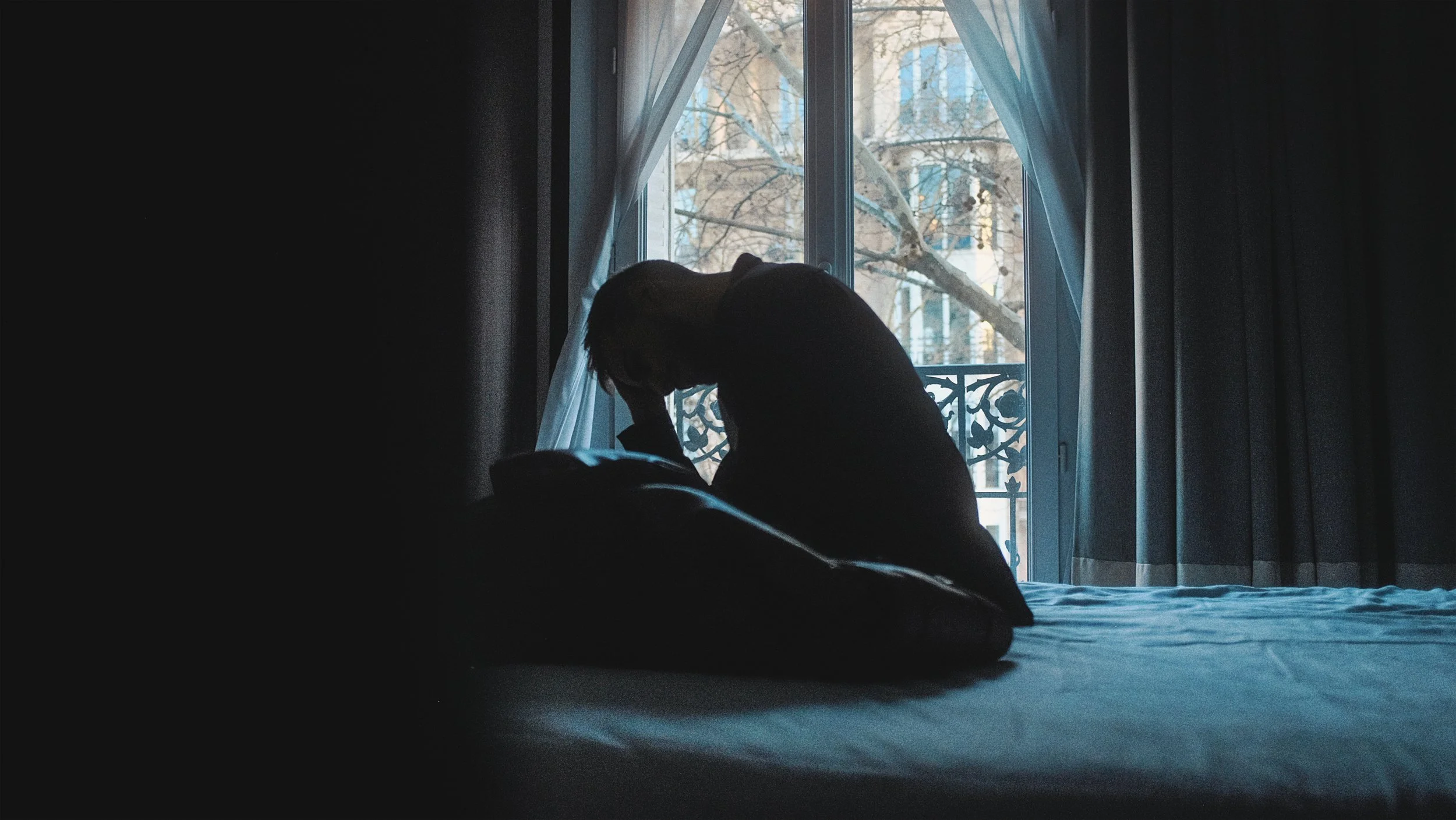 Une personne assise sur un lit près d'une fenêtre, en train de se tenir la tête dans les mains, silhouette sombre dans une pièce peu éclairée, avec un balcon et des arbres en arrière-plan.