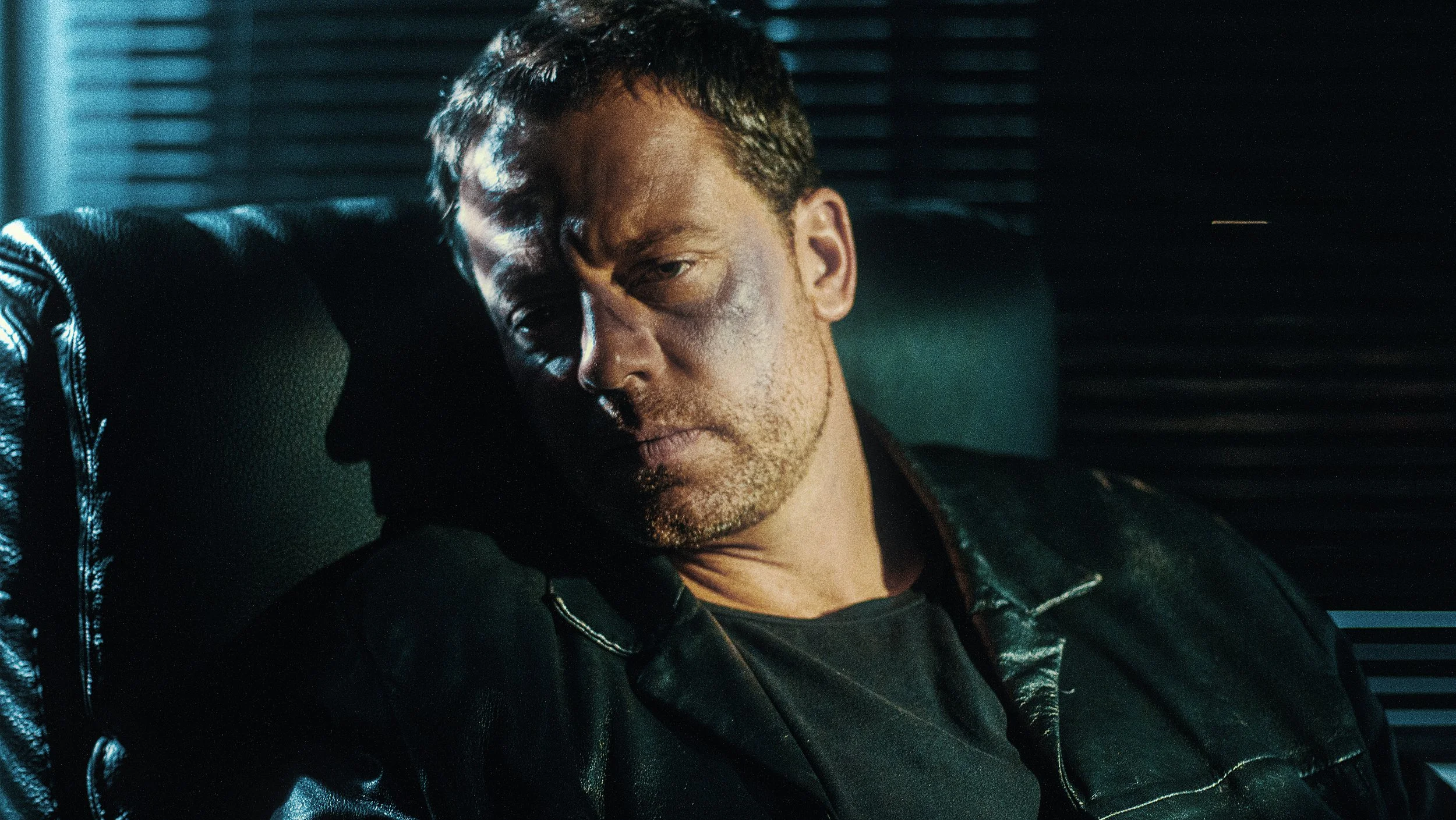 Un homme avec un visage marqué, s'appuyant contre un fauteuil en cuir, dans un environnement sombre et industriel.