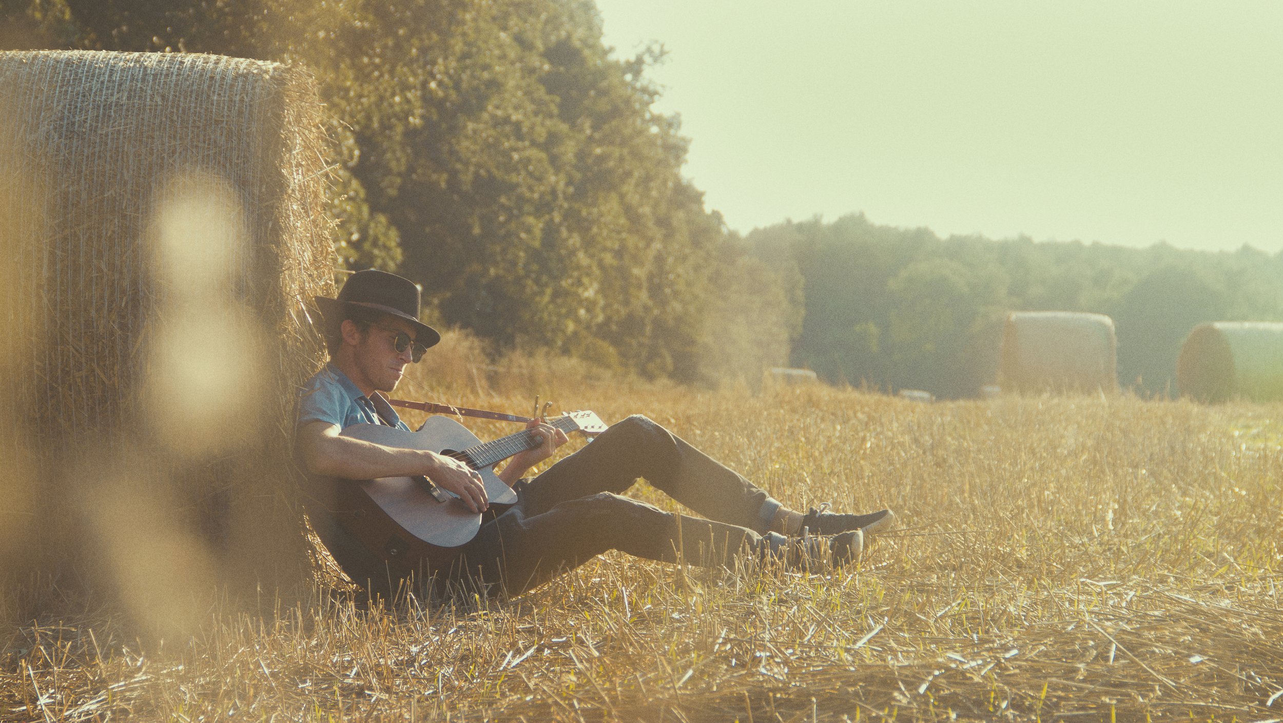 Homme jouant de la guitare dans un champ lors d'une journée ensoleillée, s'appuyant contre une botte de foin.