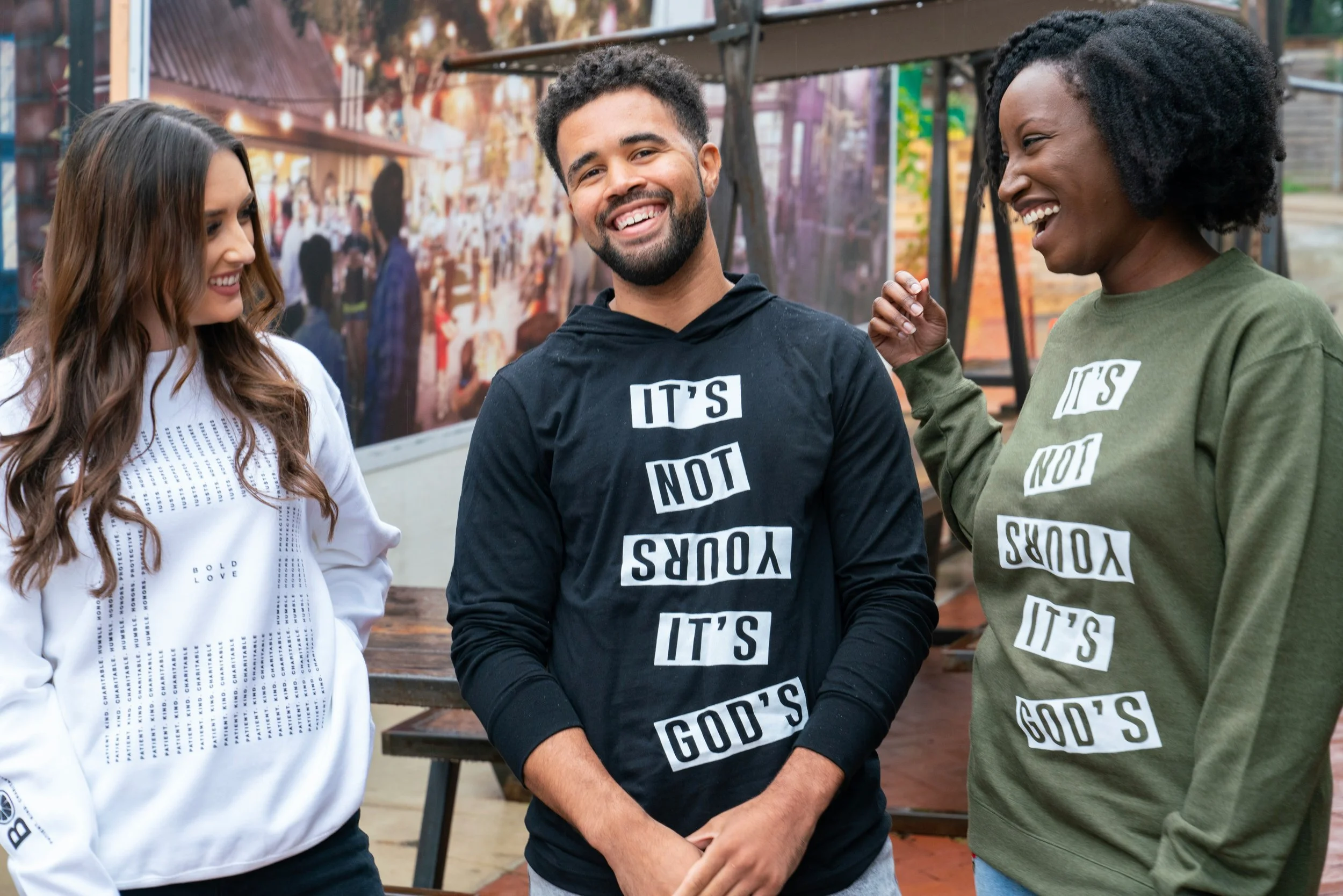 Three friends standing outdoors, smiling and talking. The man in the middle wears a black hoodie and the women on each side wear a white and green sweatshirt, with positive messages. They appear happy and engaged in conversation.