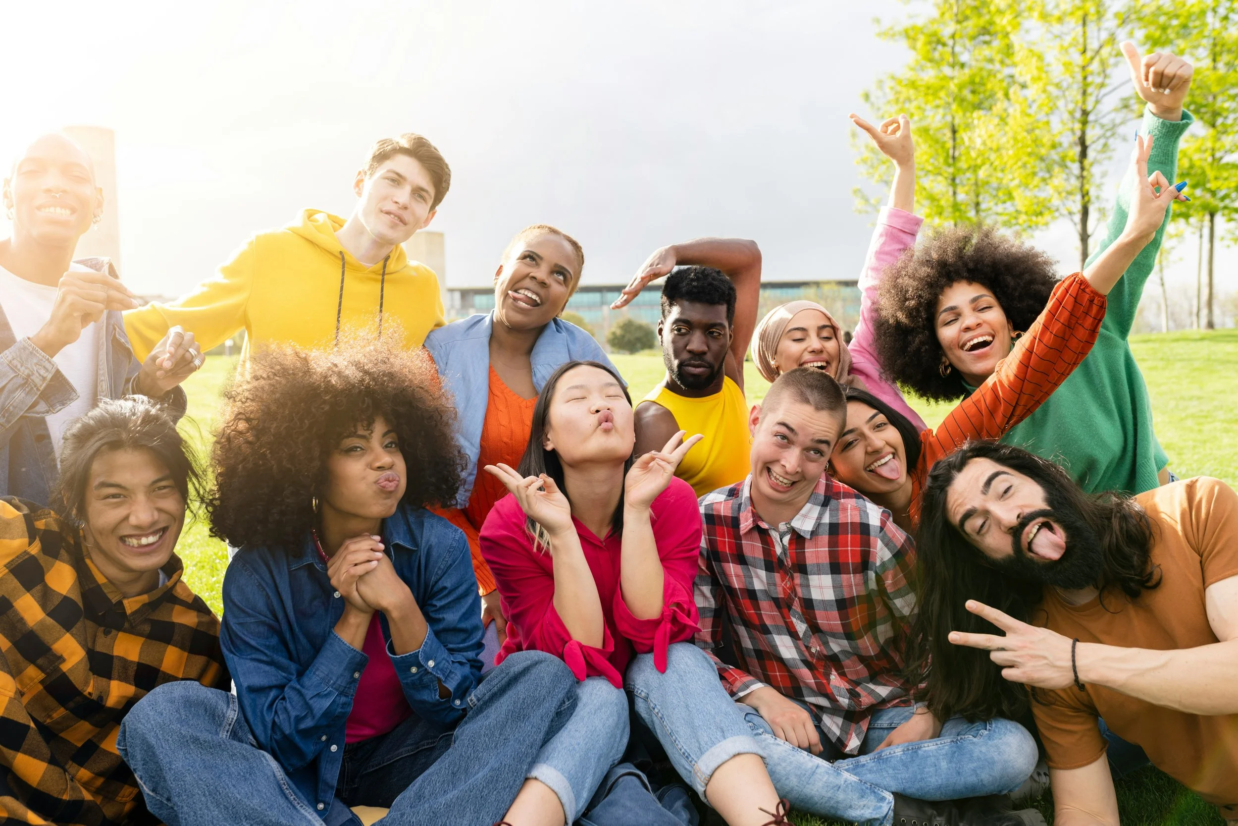 A diverse group of young adults outdoors in a park, smiling and making playful gestures.