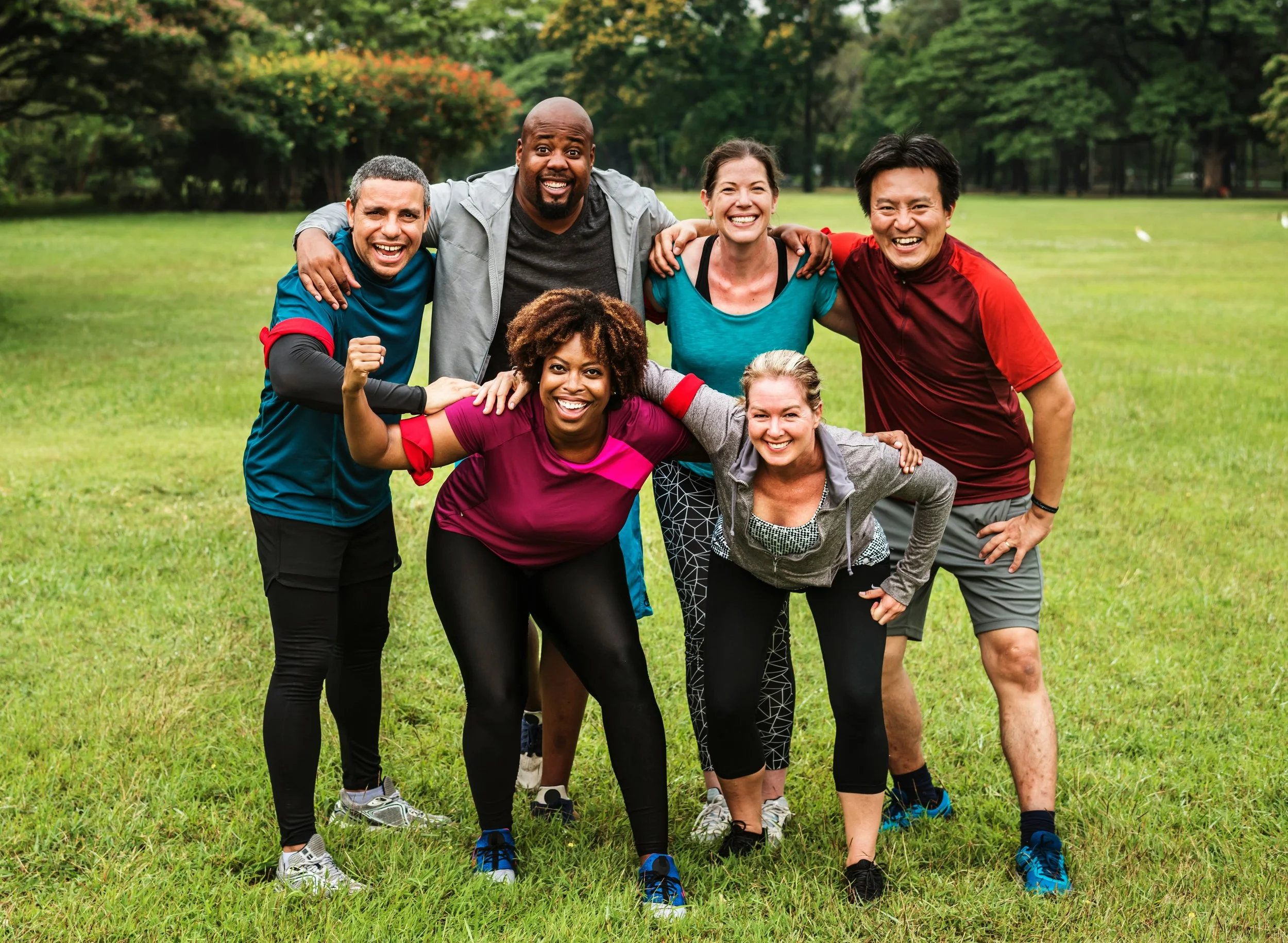 Group of seven diverse friends in athletic clothing smiling and posing together in a park on a grassy field.