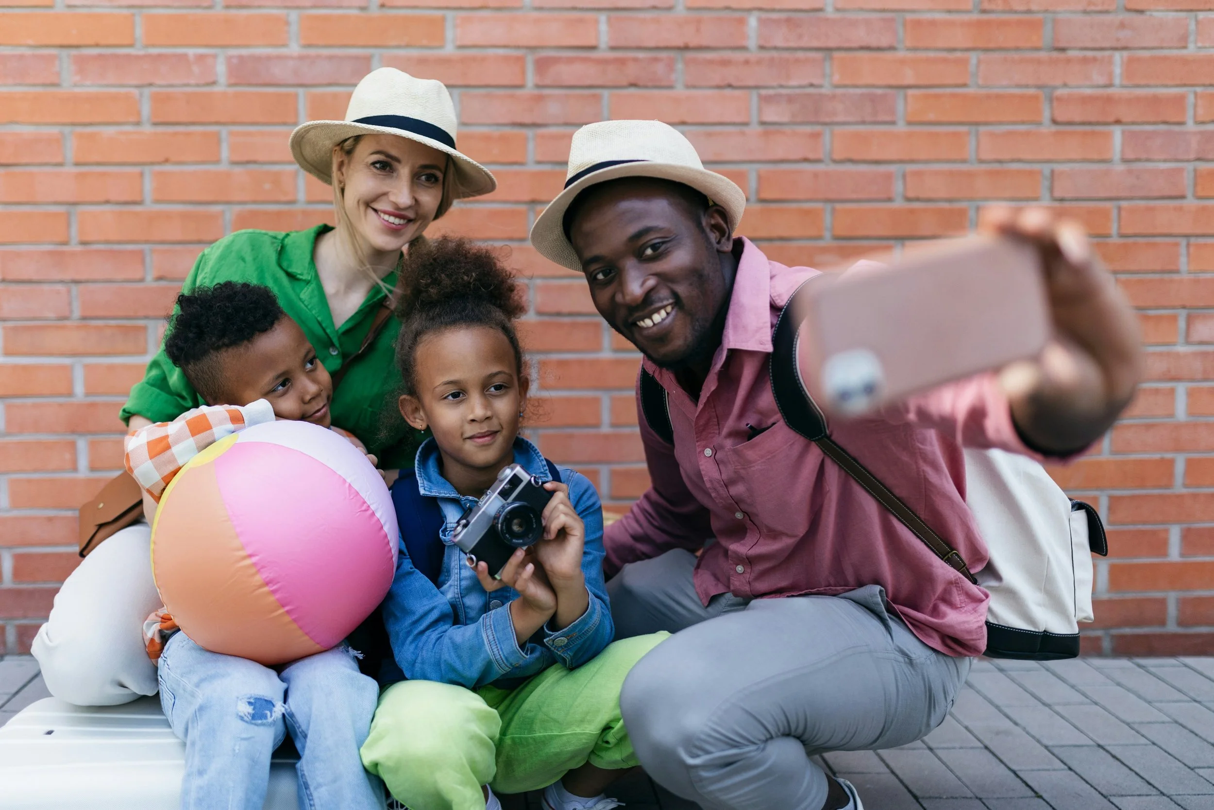 A diverse group of four children and two adults taking a selfie outdoors in front of a brick wall. Some children are holding cameras and a beach ball.