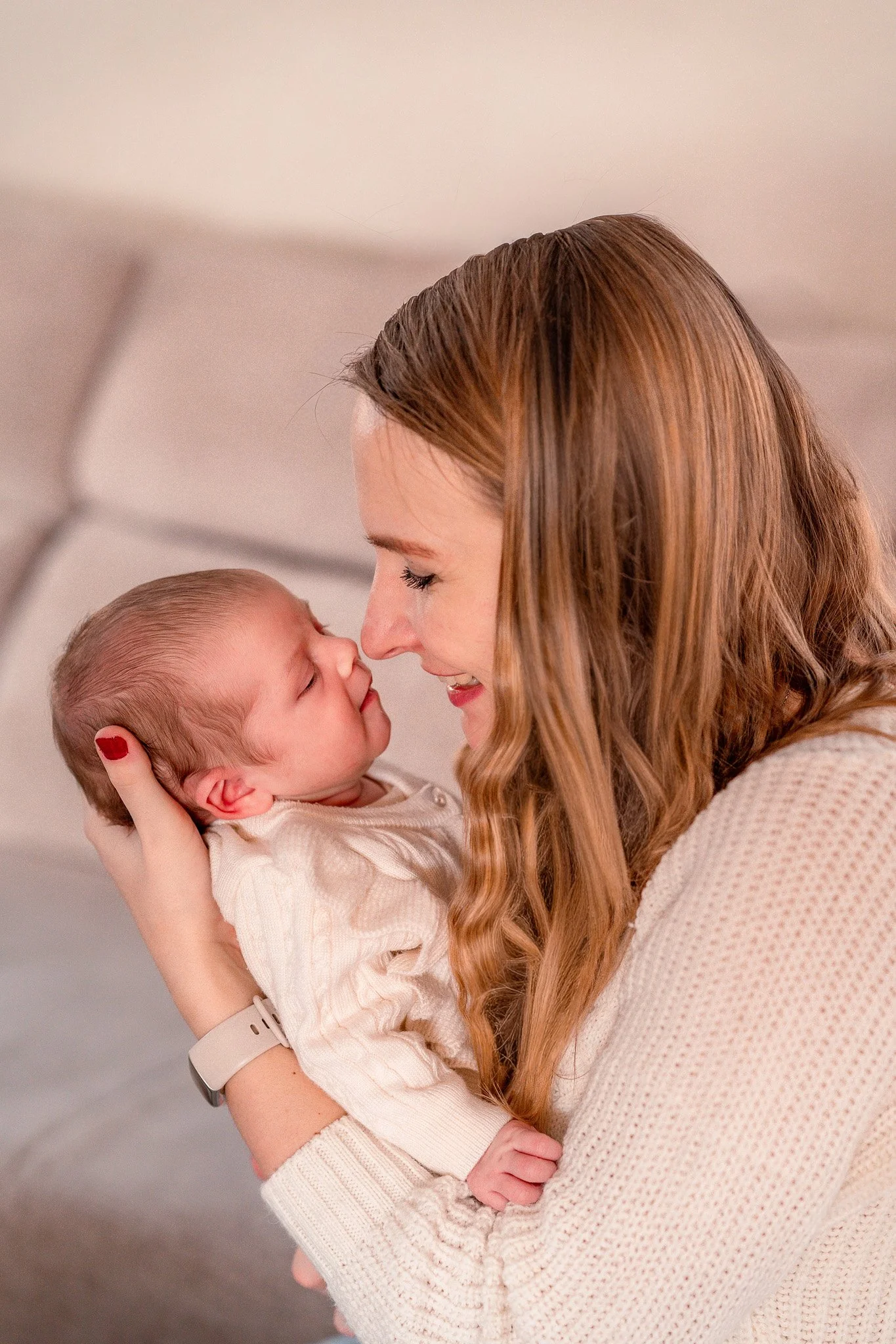 A woman with red hair and a beige sweater holding a sleeping baby with a cream-colored sweater, touching noses in a close-up moment of affection.