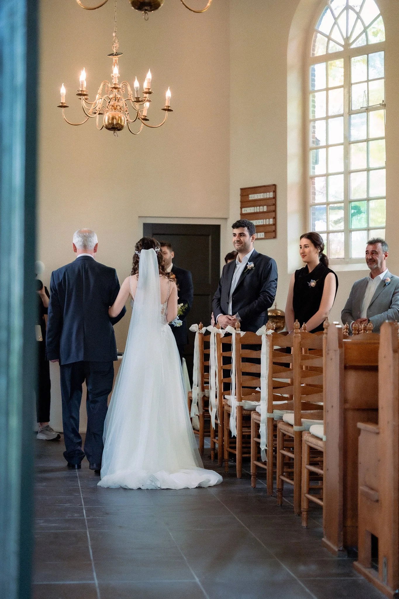 A bride and groom standing with officiant at a wedding inside a church, with guests seated and a large arched window behind them.