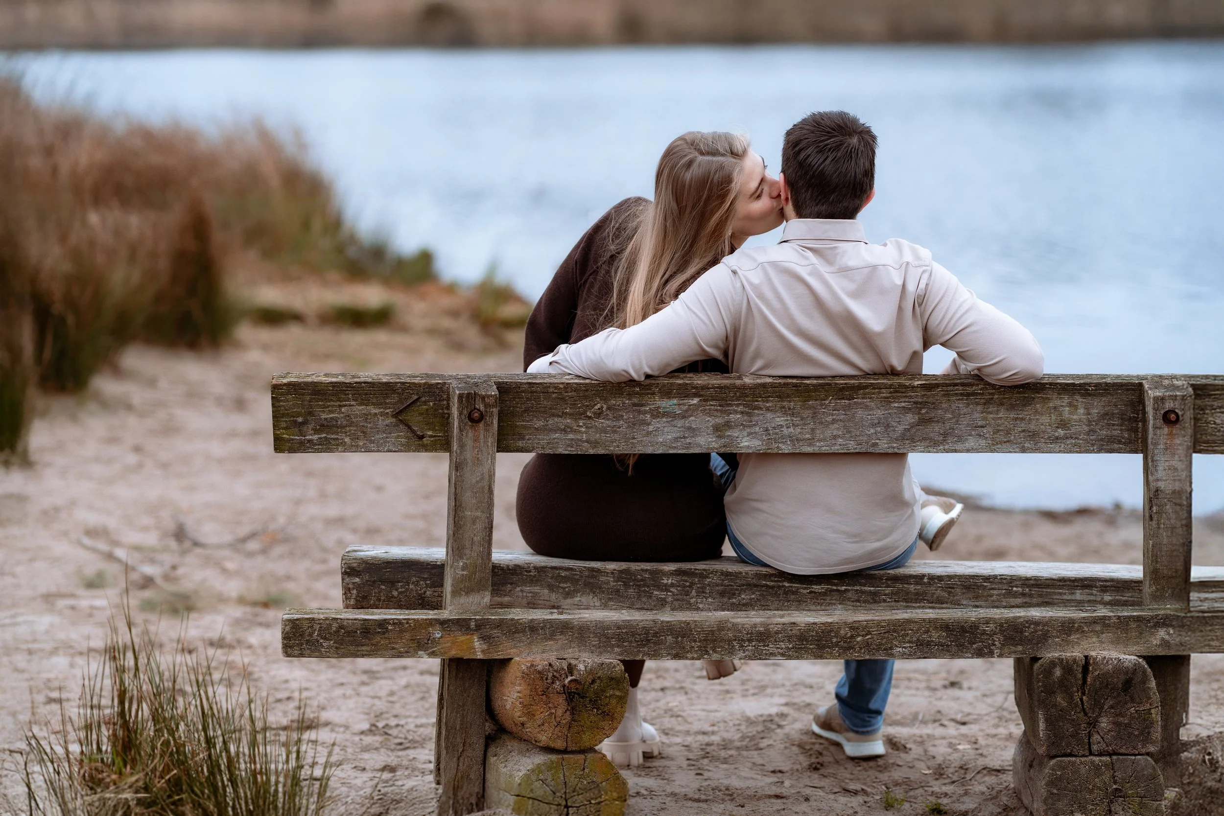 A couple sitting on a wooden bench by the water, sharing a kiss.