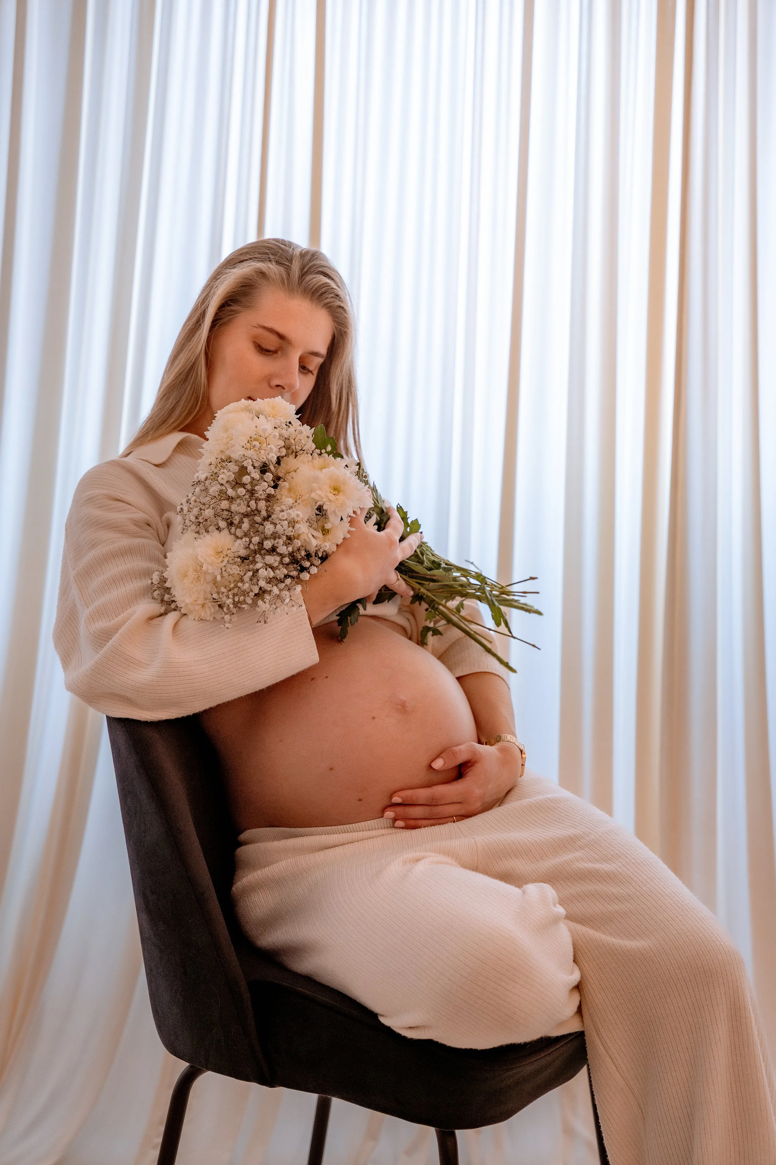 Pregnant woman sitting on a chair, holding a bouquet of white and light pink flowers, with her hand on her belly, in front of light-colored curtains.