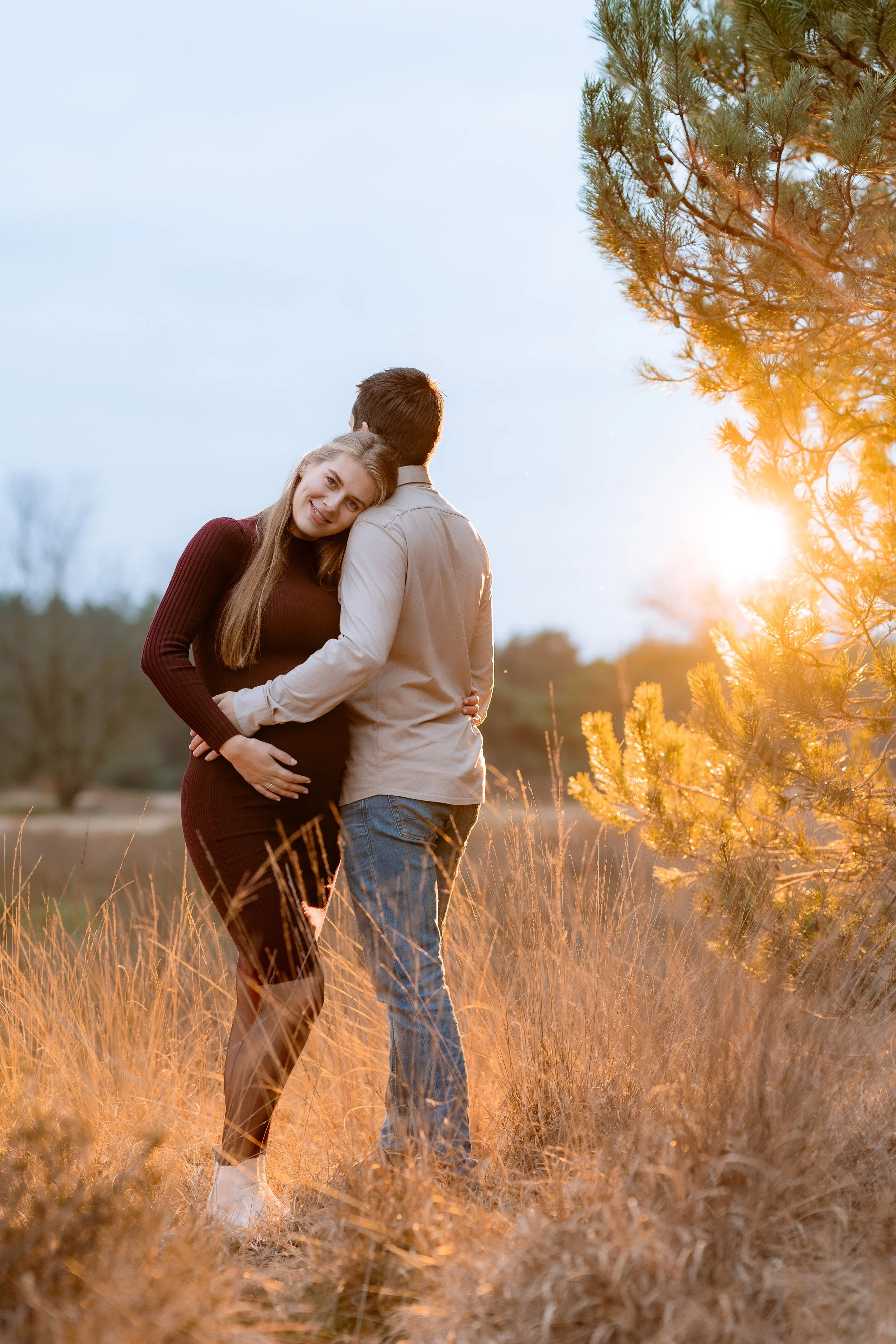 A couple standing in a field during sunset, with the woman resting her head on the man's shoulder, both facing away from the camera.