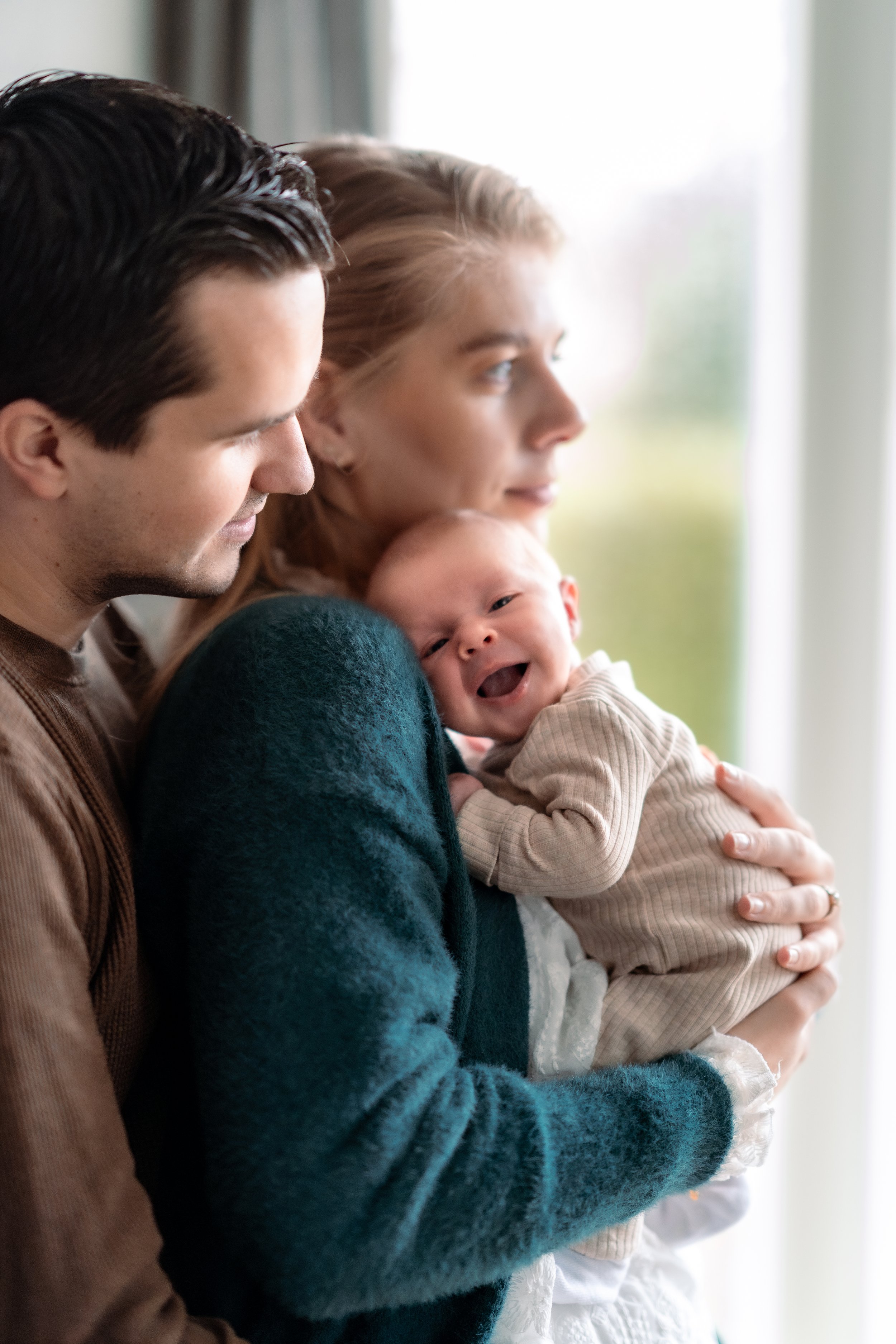 A family of three, a man, woman, and baby, standing near a window. The woman is holding the smiling baby, and they are all bundled in warm clothes with soft expressions.
