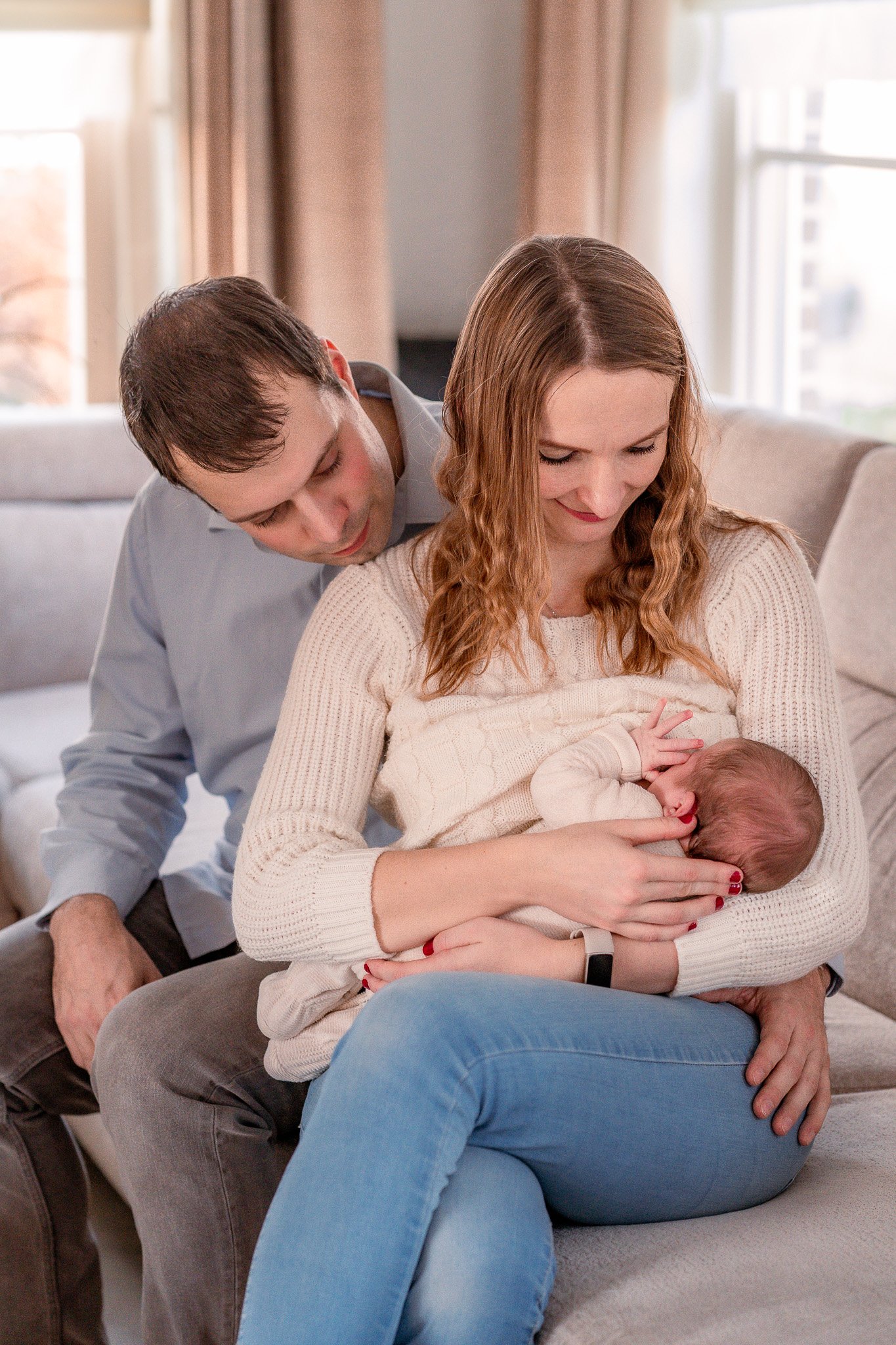 A woman breastfeeding a baby, with a man leaning over and looking at them, in a cozy living room with large windows and beige curtains.