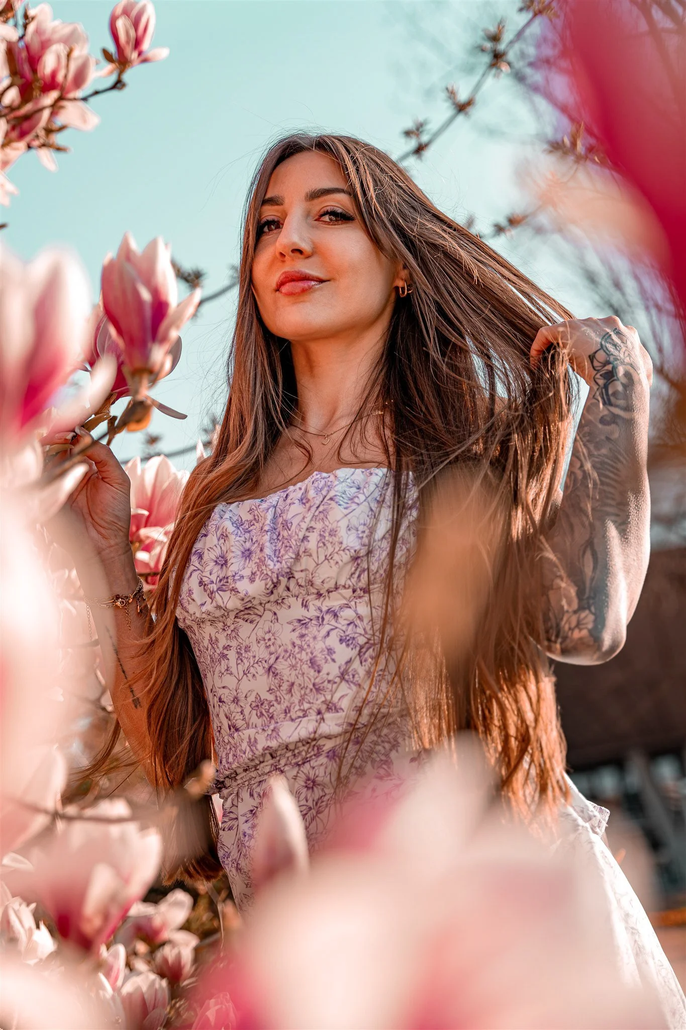 A young woman with long brown hair standing among blooming pink magnolia flowers during daytime, wearing a floral dress and holding a lock of hair, looking at the camera with a confident expression.