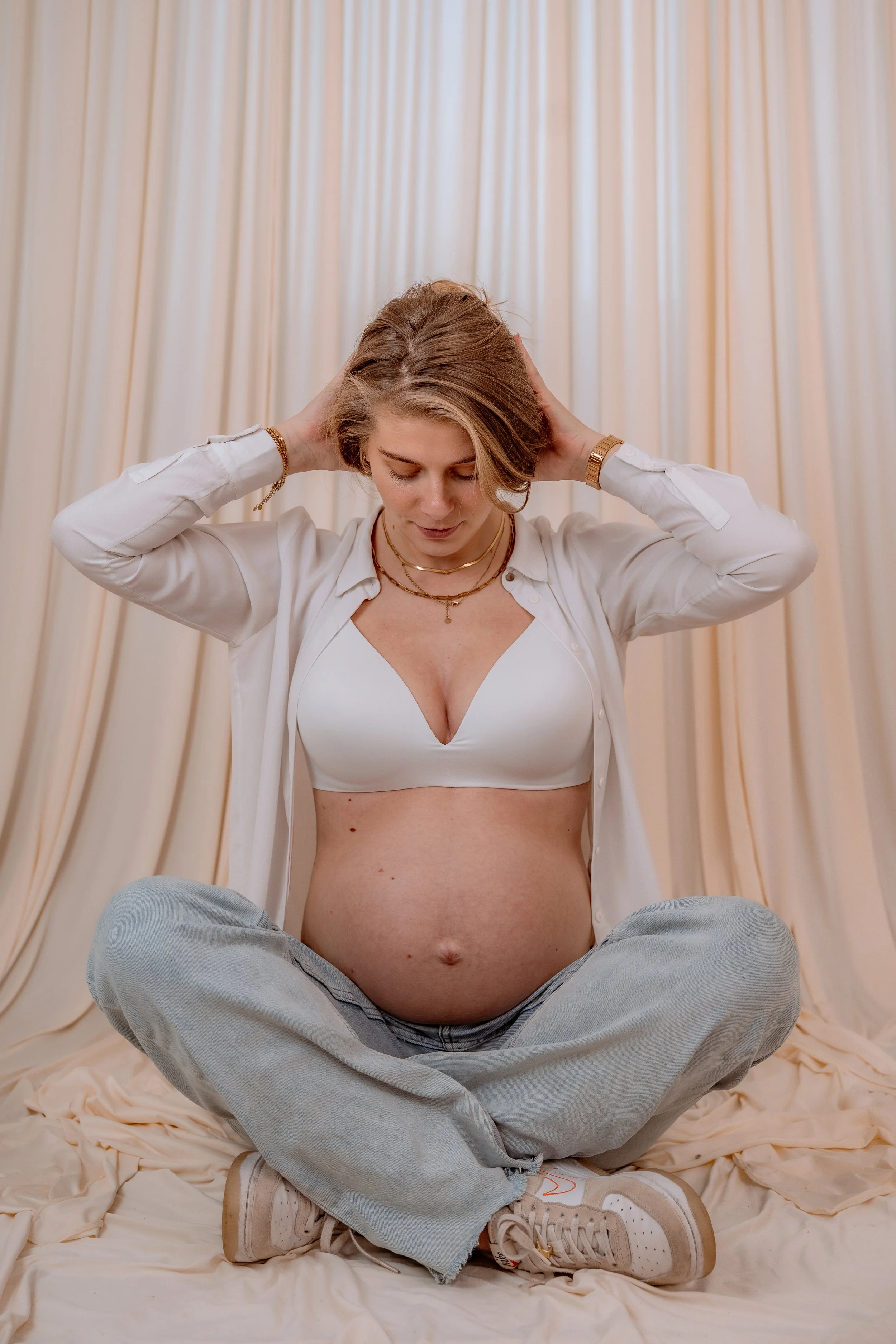 A pregnant woman sitting cross-legged on a cream-colored cloth, wearing a white bra, light blue jeans, and sneakers, with a white shirt open and draped loosely. She has short blonde hair, multiple necklaces, a watch, and a bracelet, and holds her han