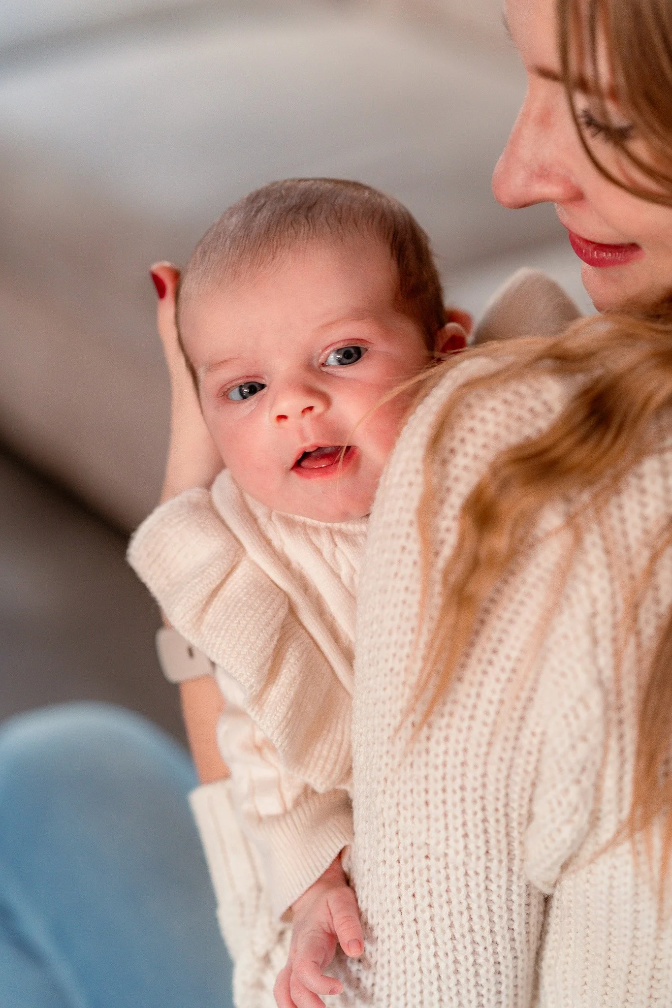 A young woman holding a baby girl with blue eyes and light skin, both wearing cream-colored sweaters.
