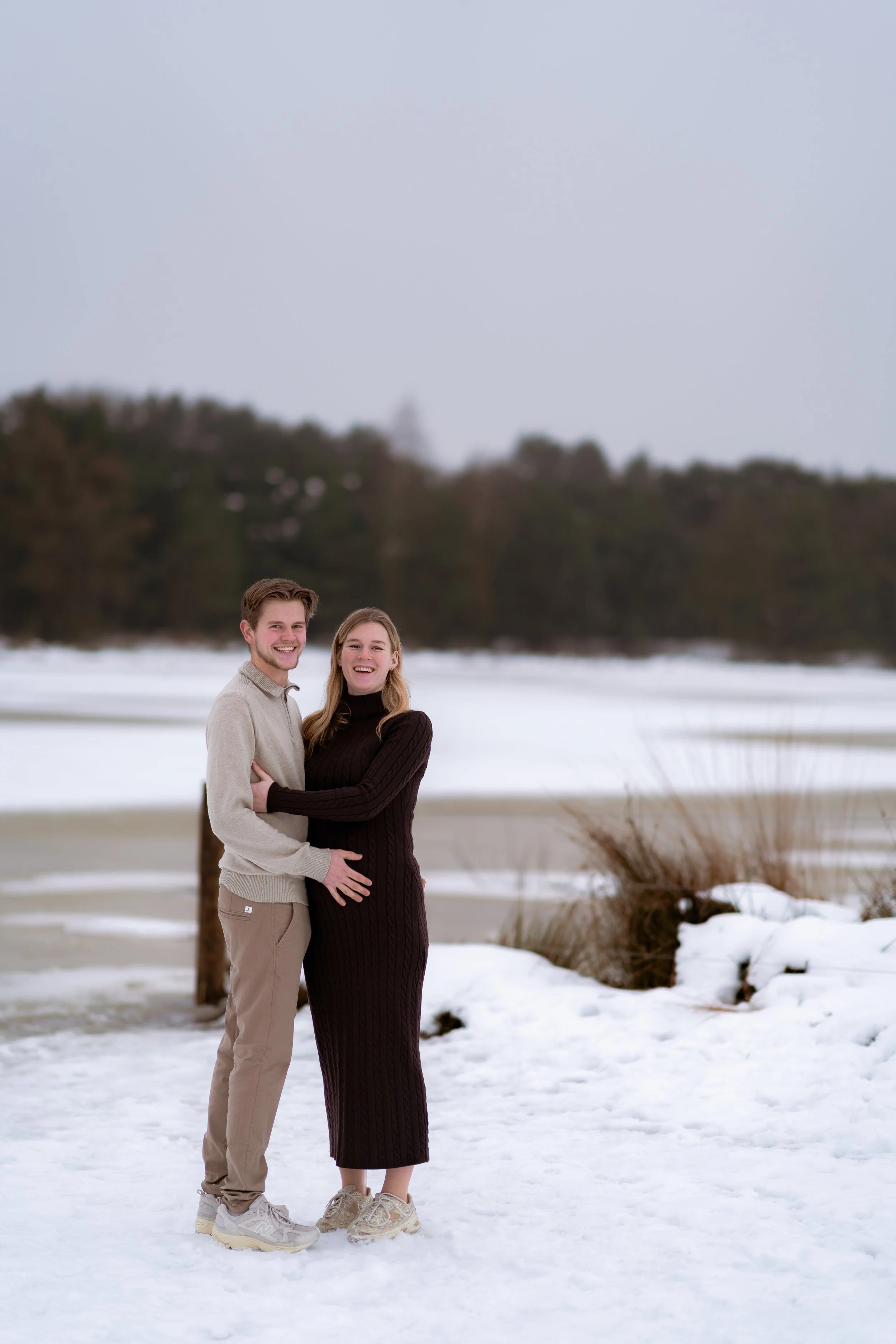A couple stands on snow-covered ground in winter, smiling at the camera with a frozen lake and trees in the background.