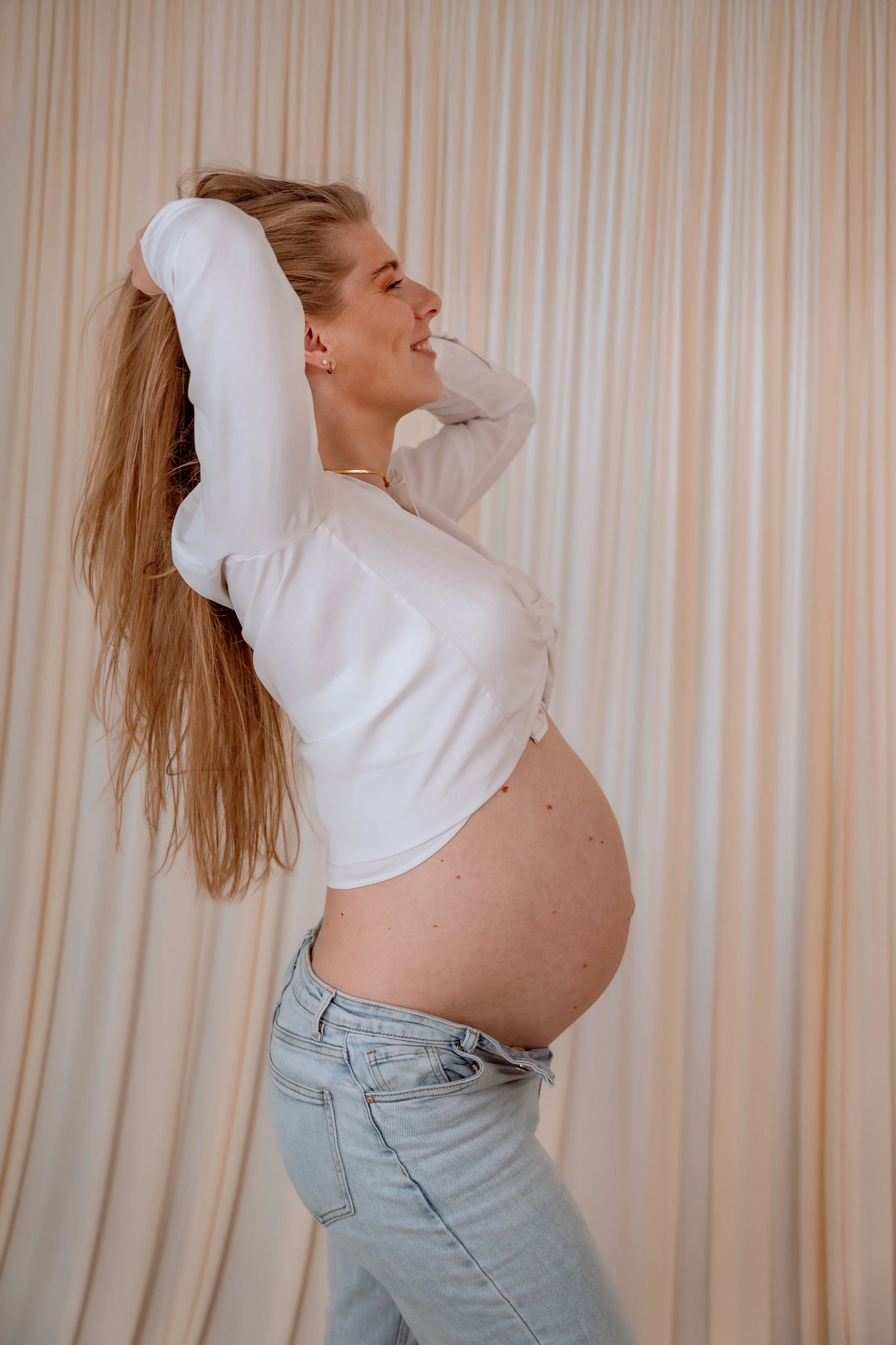 Pregnant woman smiling with hands behind her head, wearing a white blouse and light blue jeans, standing in front of beige curtains.