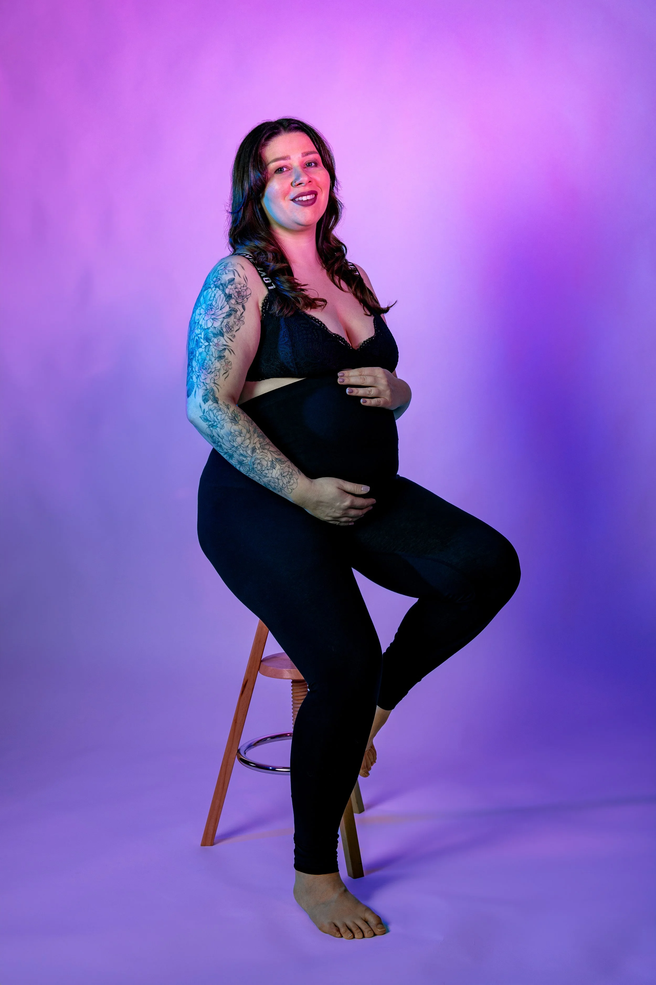 A pregnant woman with tattoos on her arm, seated on a stool, smiling, and holding her belly, against a colorful purple and pink gradient background.