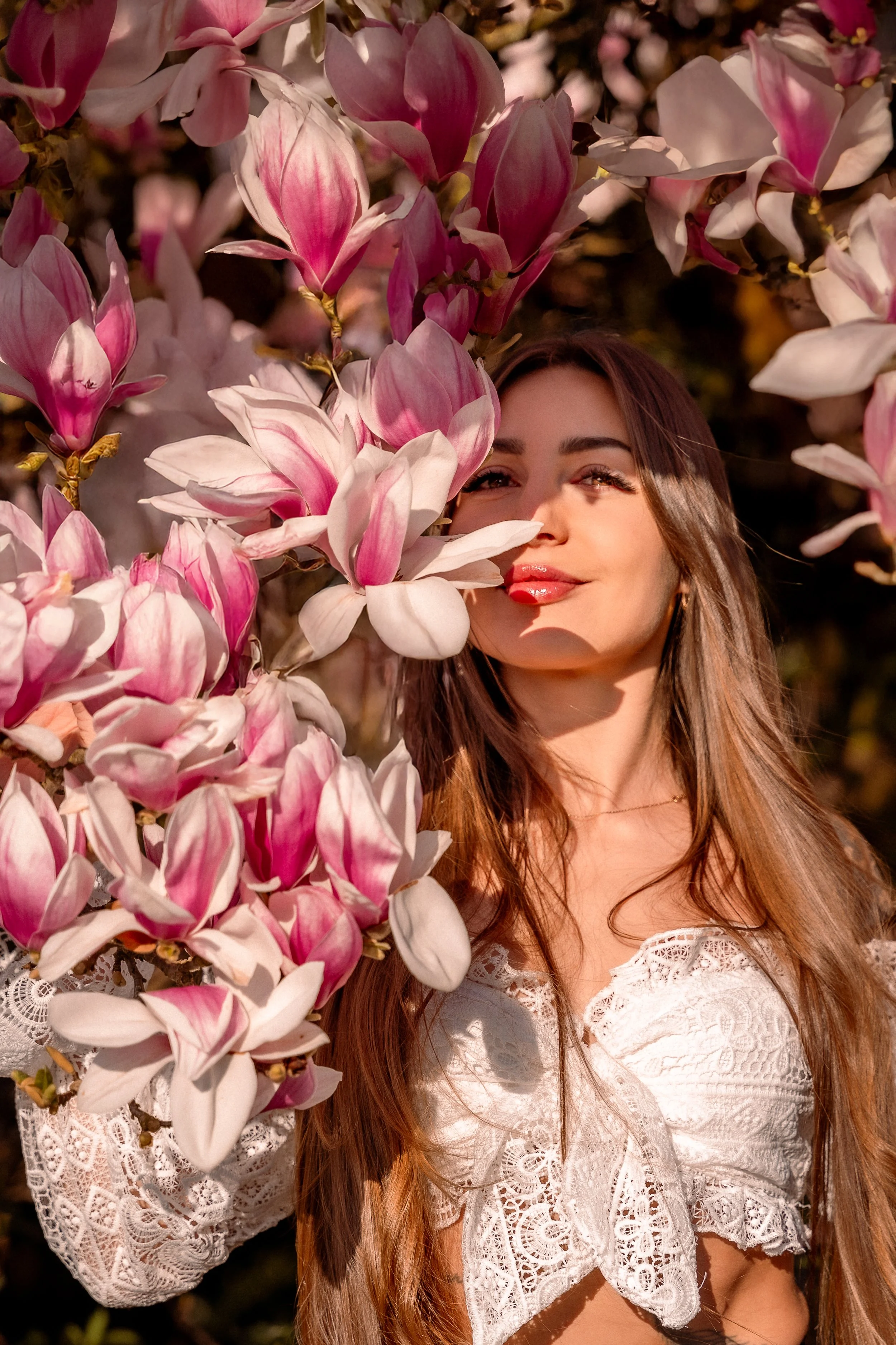A woman with long brown hair, wearing a white lace top, standing among pink and white magnolia flowers, partially covering her face, with sunlight illuminating her and the flowers.