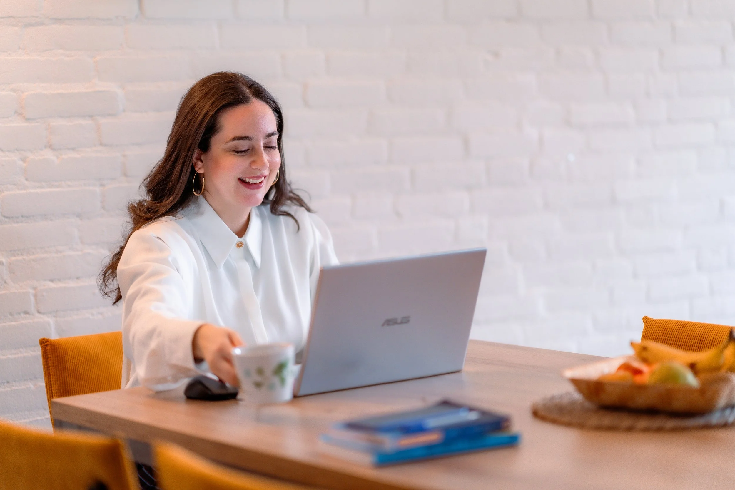 A woman with brown hair and hoop earrings working on a laptop at a wooden table, smiling, with a white brick wall in the background, a mug in her hand, and a bowl of fruit on the table.