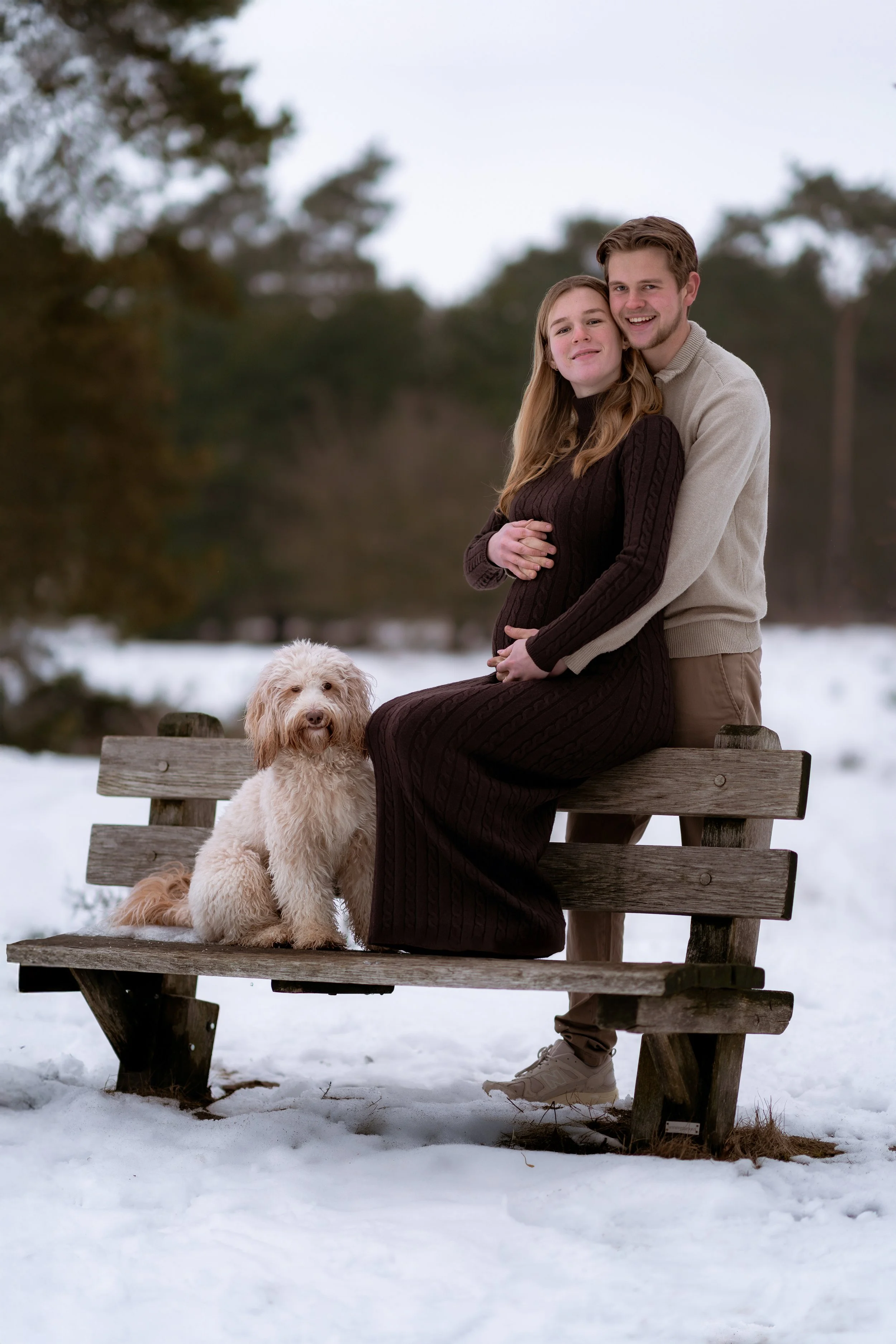 A couple and their dog sitting on a wooden park bench in a snowy landscape during winter. The woman is pregnant and sitting on the bench, while the man is standing behind her, embracing her. The woman has long hair and is wearing a dark brown dress. 