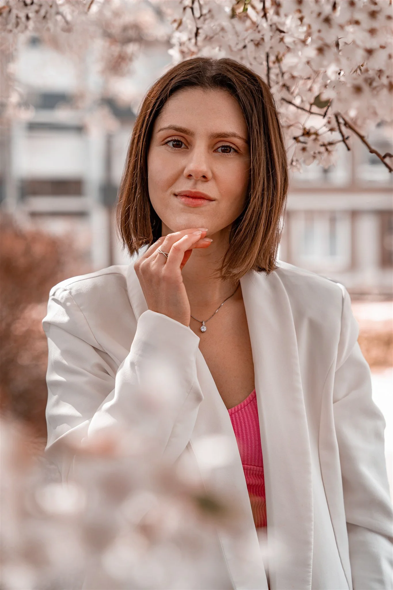 A woman with short brown hair wearing a white blazer and pink top, standing outdoors near blooming cherry blossom trees.