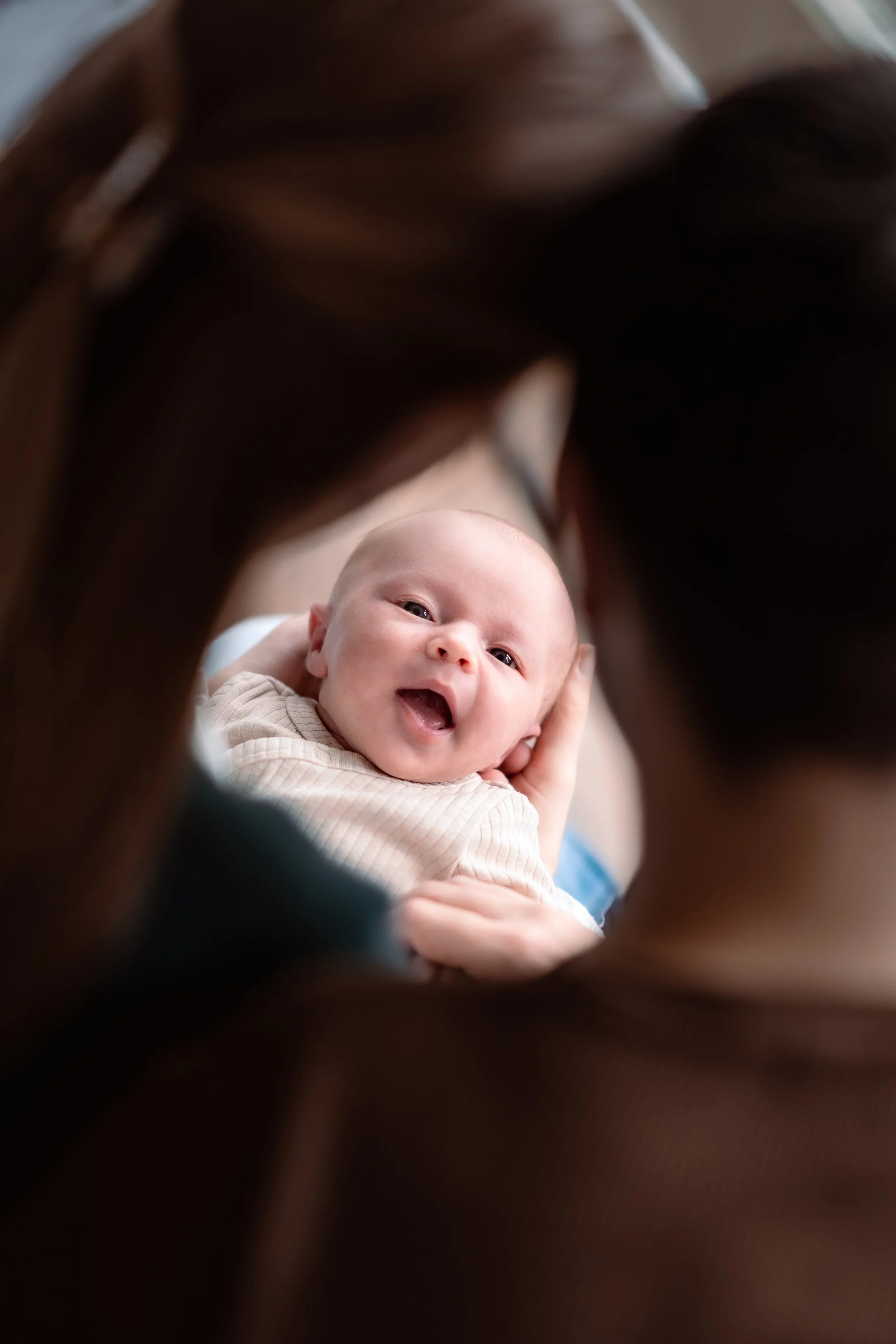 A baby smiling and looking up, held by an adult, with the photo taken through a frame of hair.