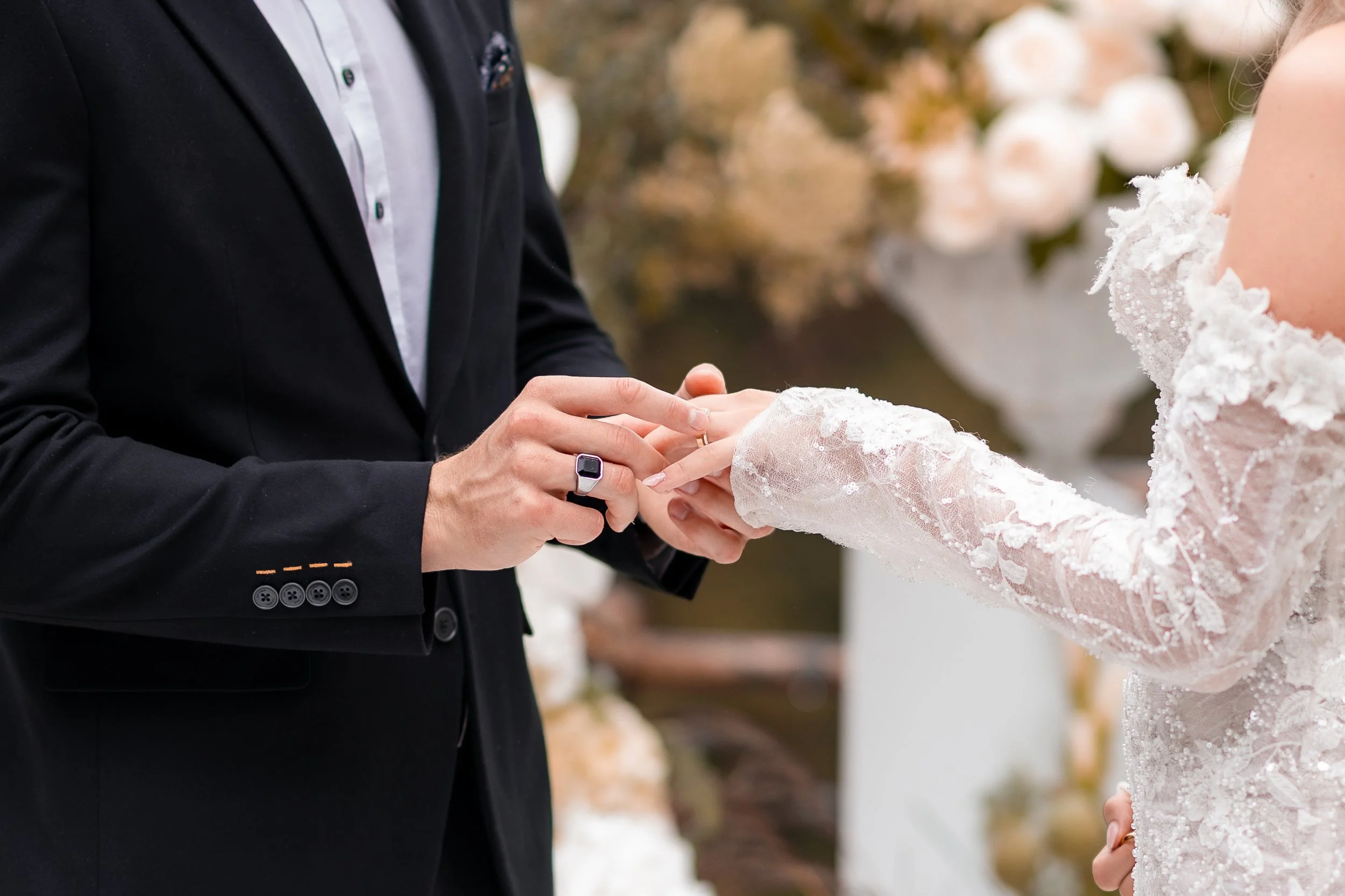 Close-up of a wedding ceremony with a groom placing a wedding ring on the bride's finger. The groom is dressed in a black suit, and the bride wears a white lace wedding dress. There are blurred floral decorations in the background.
