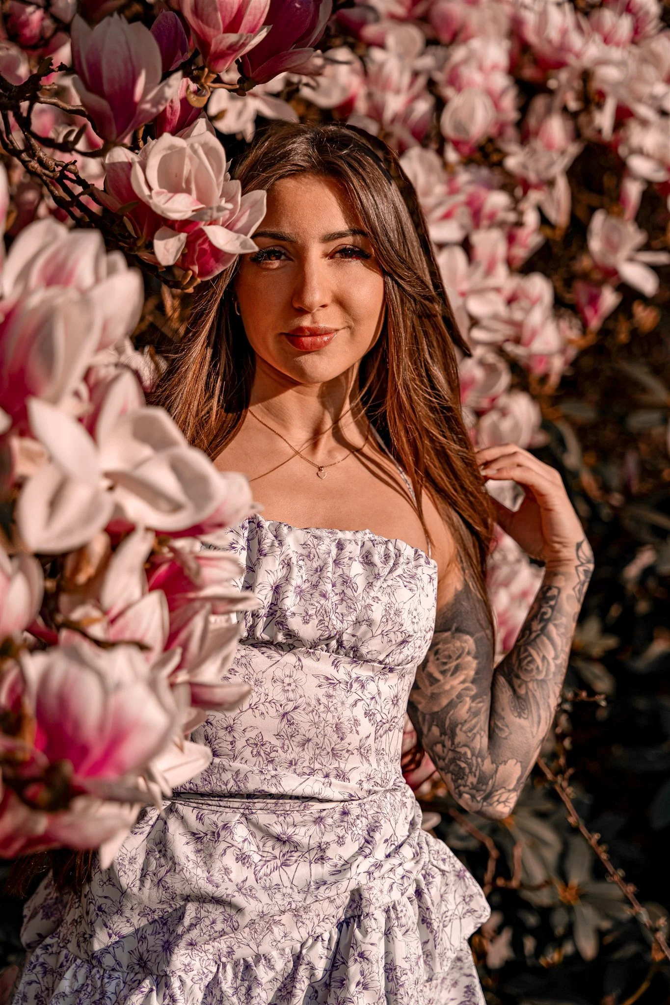 A woman with long brown hair and tattooed arm standing among pink magnolia blossoms, wearing a floral off-shoulder dress, and looking at the camera.