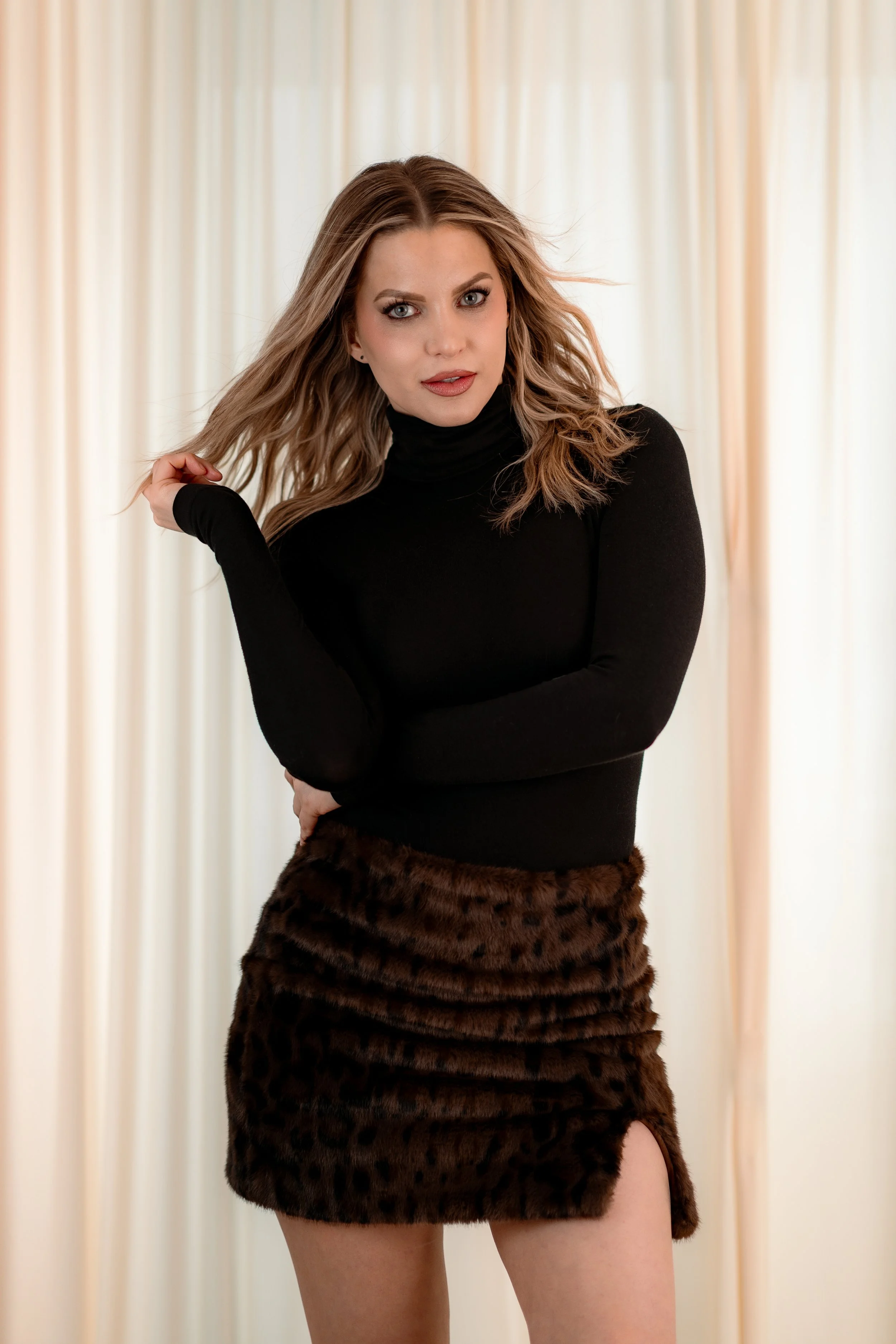 A woman with wavy blonde hair wearing a black turtleneck top and a leopard print skirt, standing in front of a light-colored curtain.