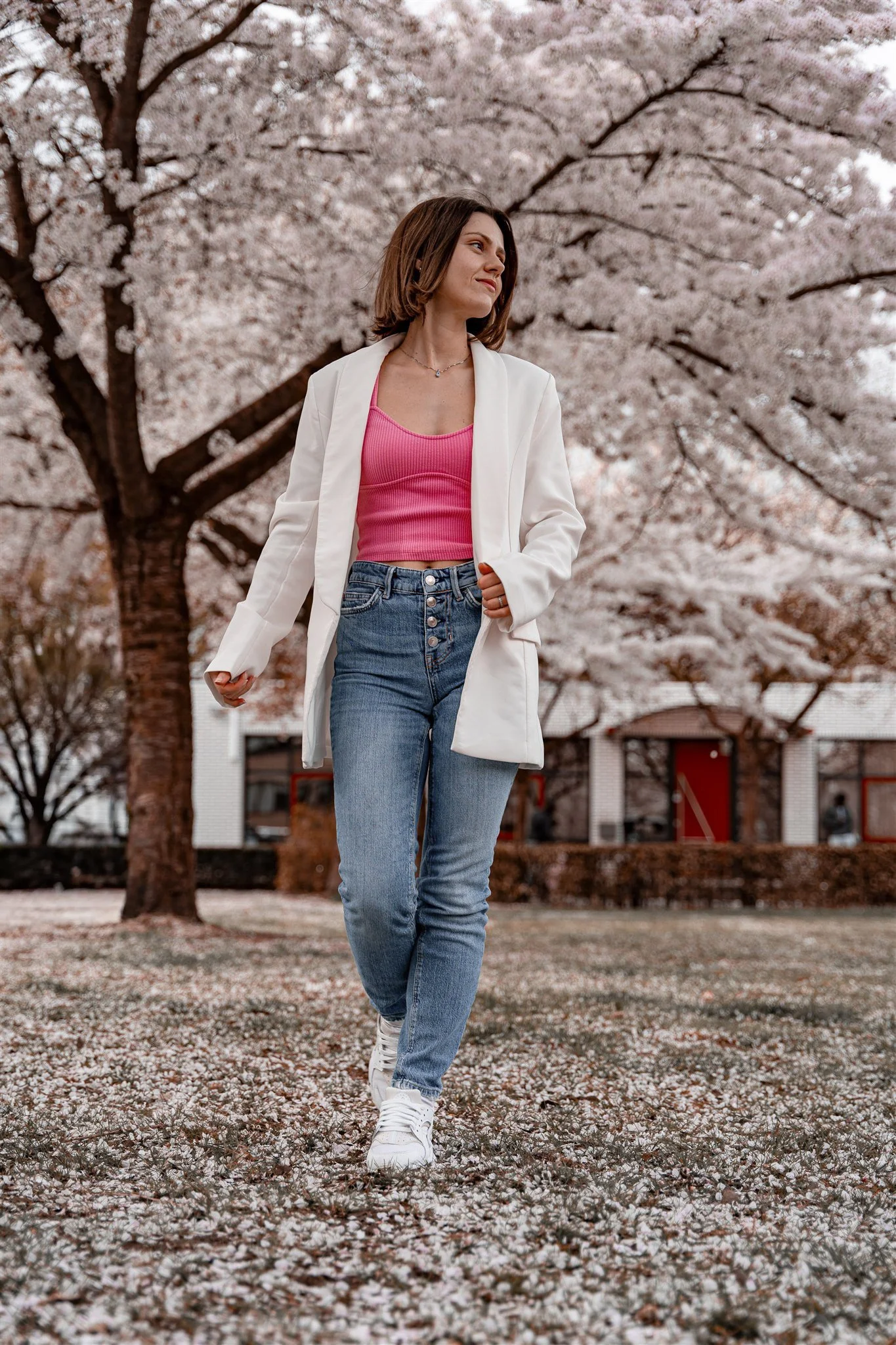 A woman walking in a park during spring with cherry blossom trees in full bloom and petals on the ground.
