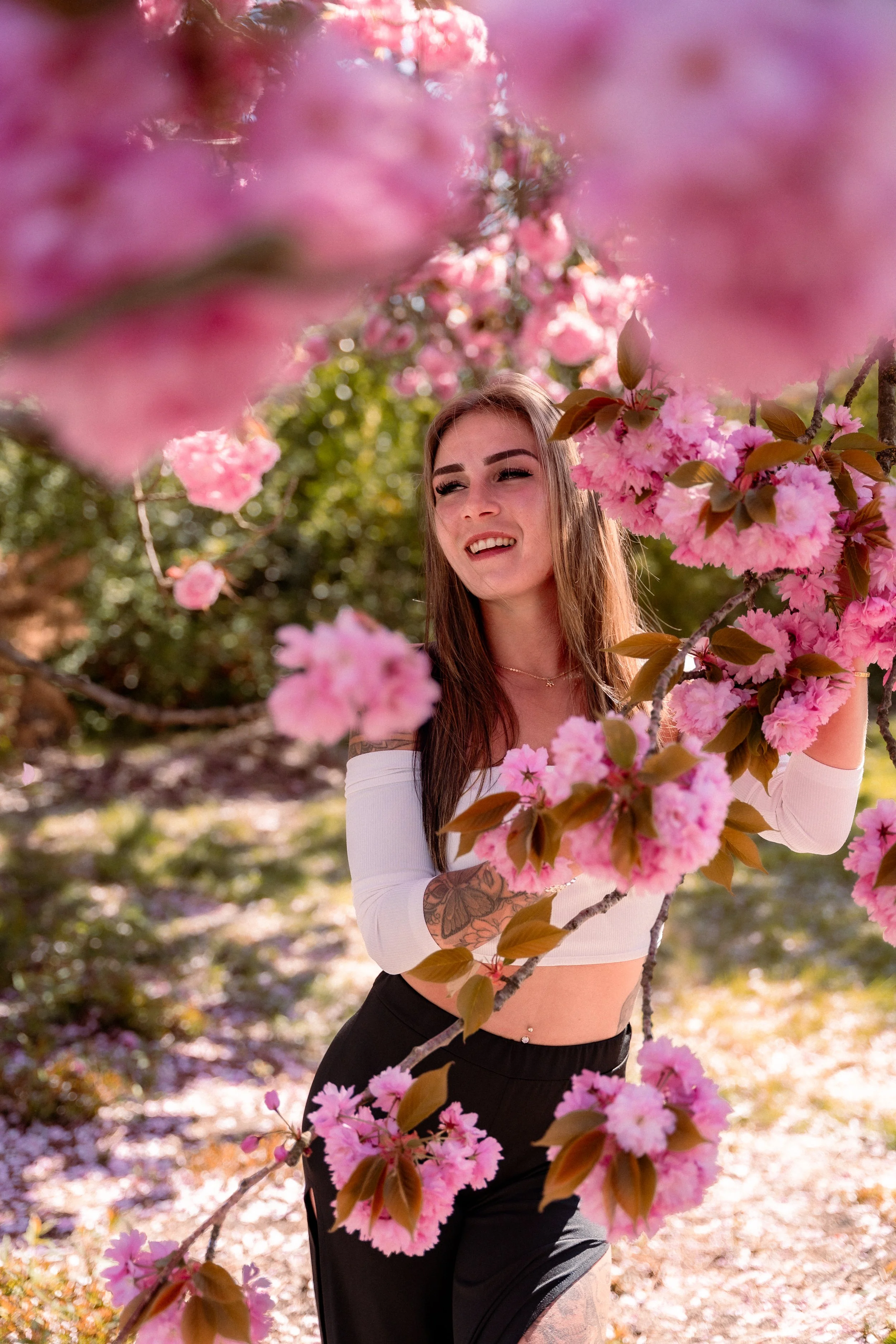 A woman is standing among pink blooming cherry blossoms outdoors, smiling and looking to the side. She has long brown hair, tattoos on her arms, a cropped white top, and black pants.