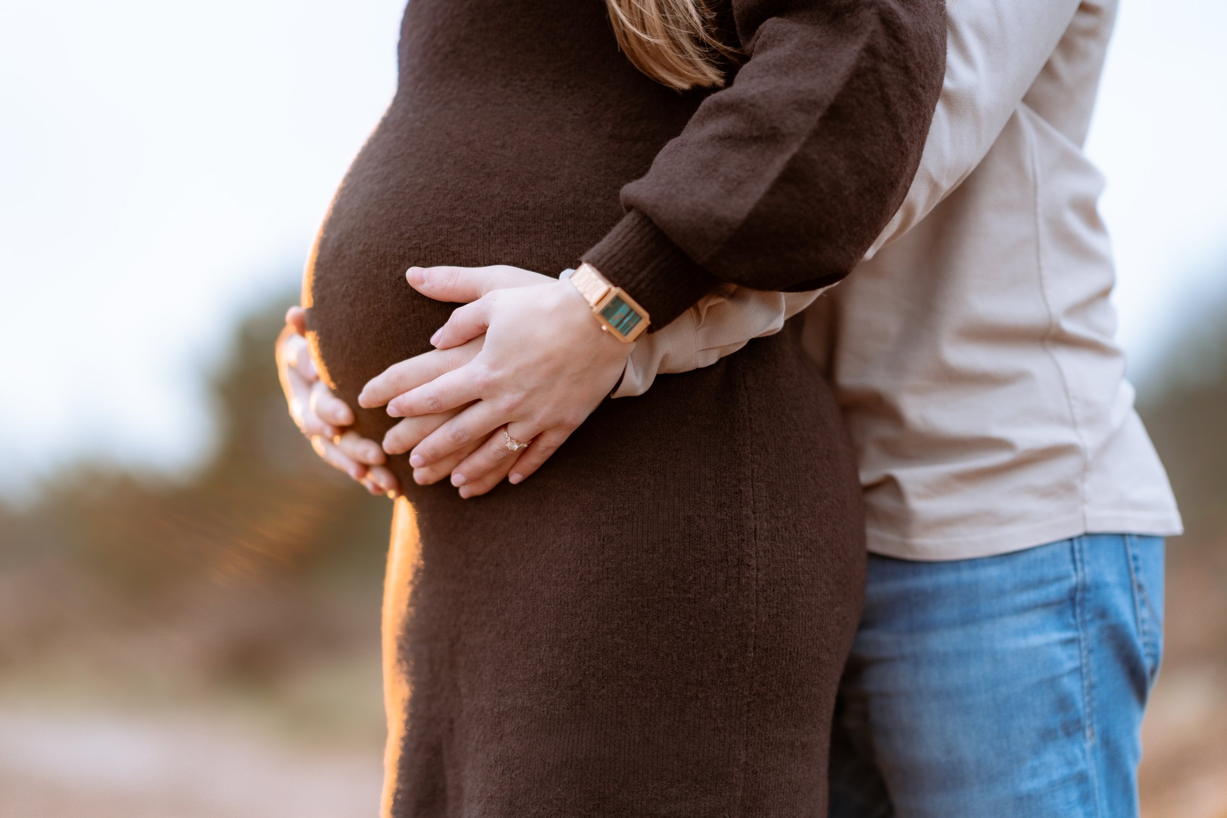 Close-up of a pregnant woman’s belly with a couple’s hands gently resting on it, outdoors during sunset.
