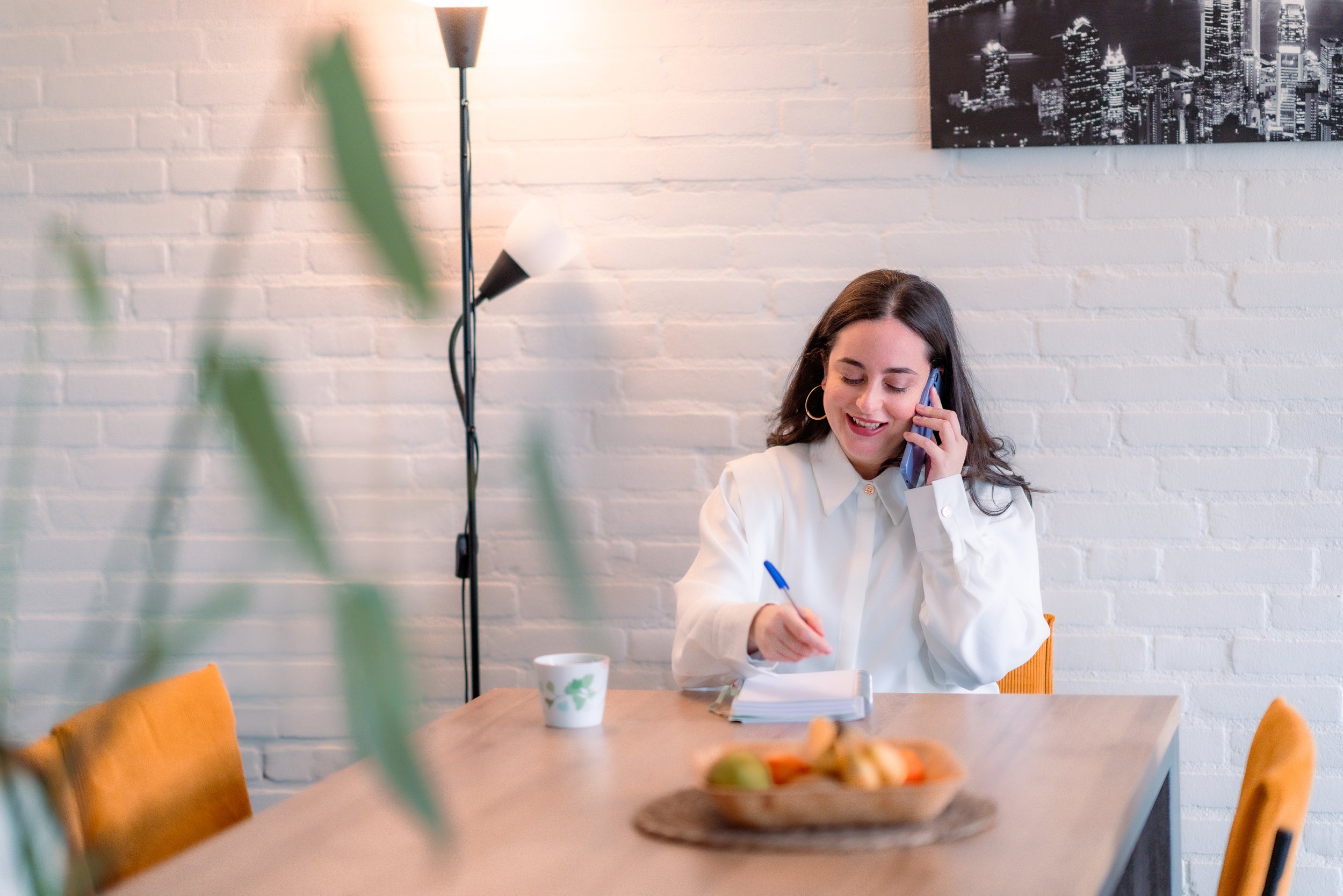 Woman with brown hair and hoop earrings, wearing a white blouse, talking on a blue cellphone while writing in a notebook at a wooden table. The table has a white cup with a green leaf design, a wooden tray with fruit, and a blue pen. Behind her is a 