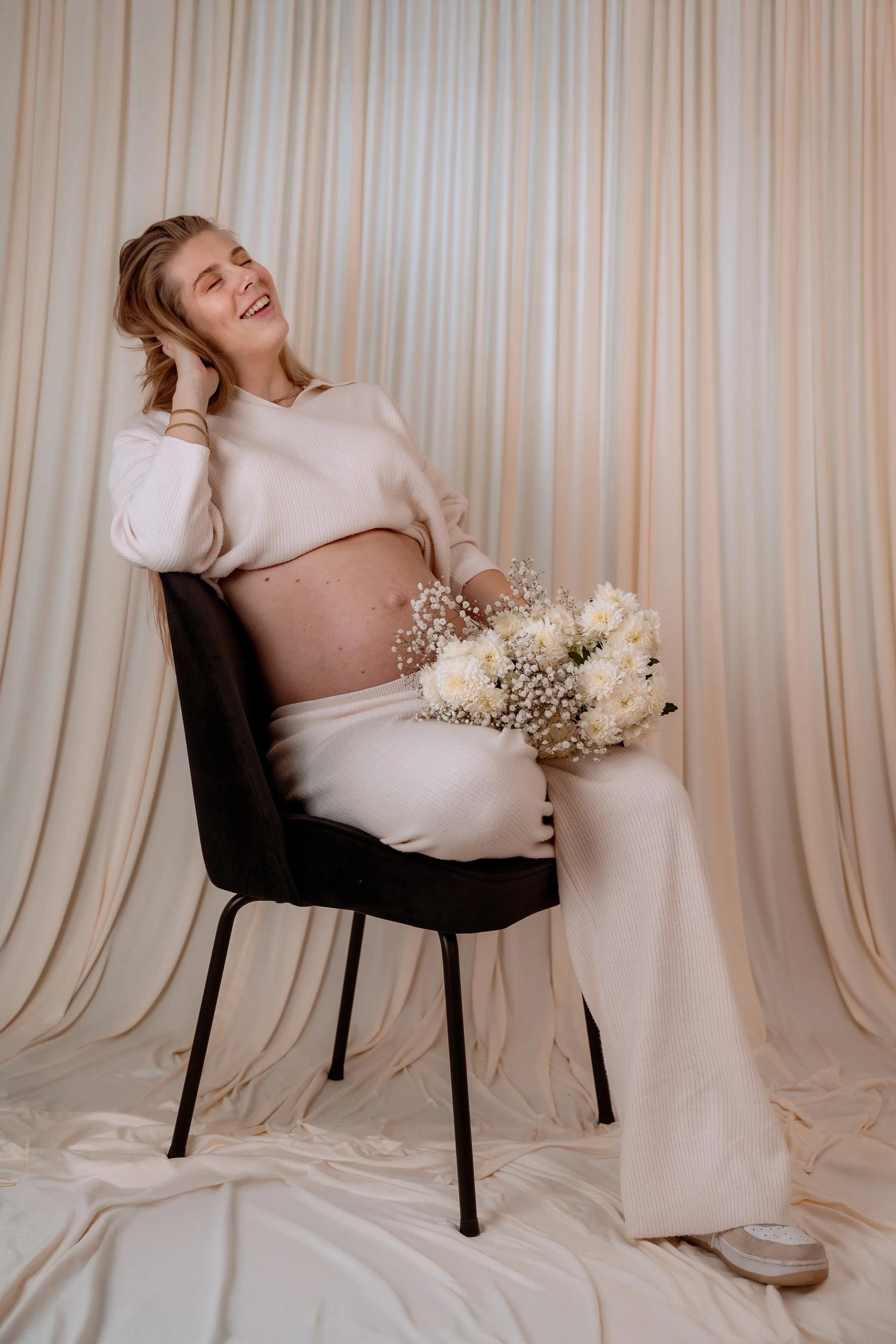 Pregnant woman sitting on a black chair, holding a bouquet of white flowers, smiling with her eyes closed, against a cream-colored curtain backdrop.