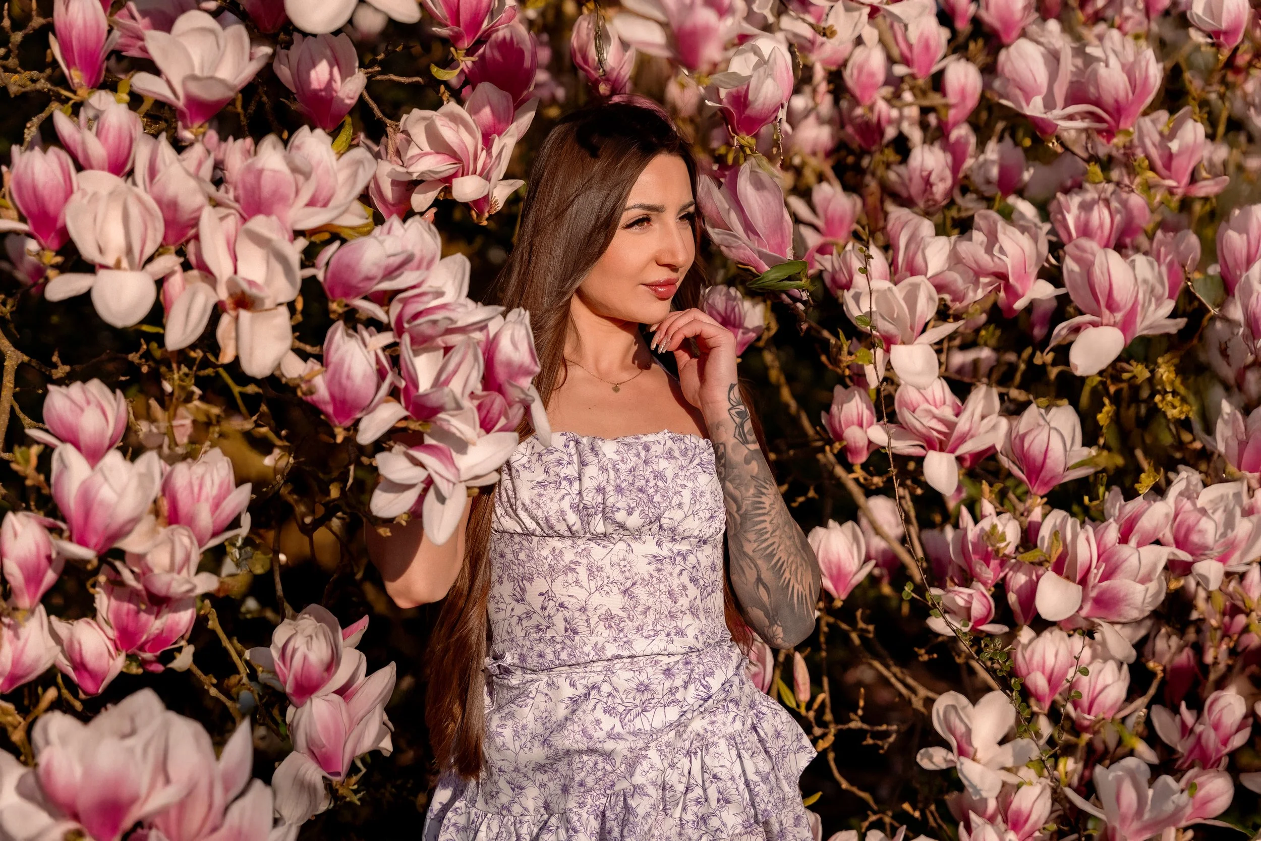 A woman with long brown hair and tattoos on her arm, wearing a white dress with purple floral patterns, stands among pink and white magnolia flowers in bloom, touching her chin with her right hand, in an outdoor setting.