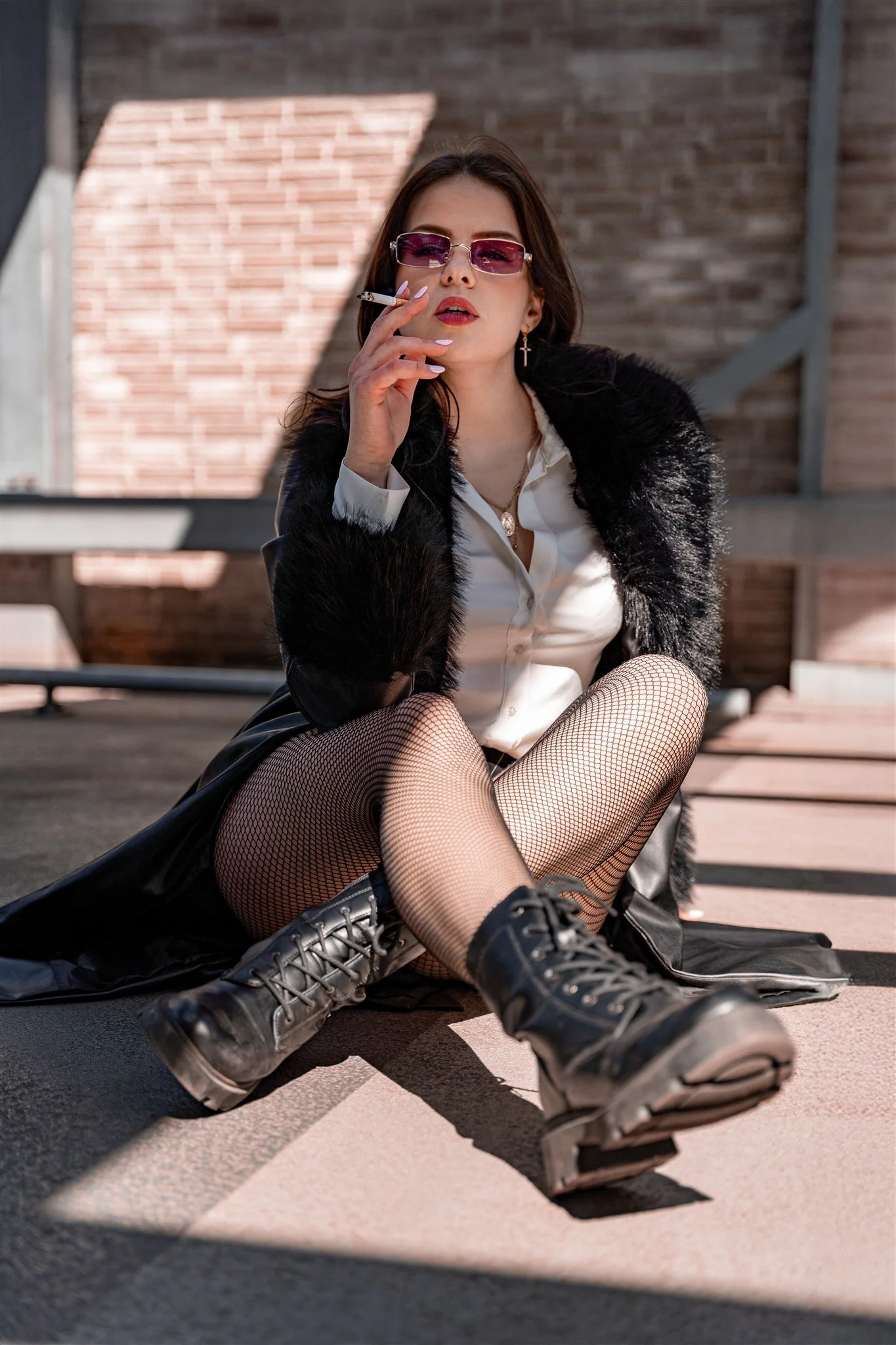A woman sitting on the ground with crossed legs, wearing fishnet stockings, black lace-up boots, a white blouse, a black jacket with fur, and purple sunglasses, holding a cigarette to her lips in an outdoor setting with brick walls.