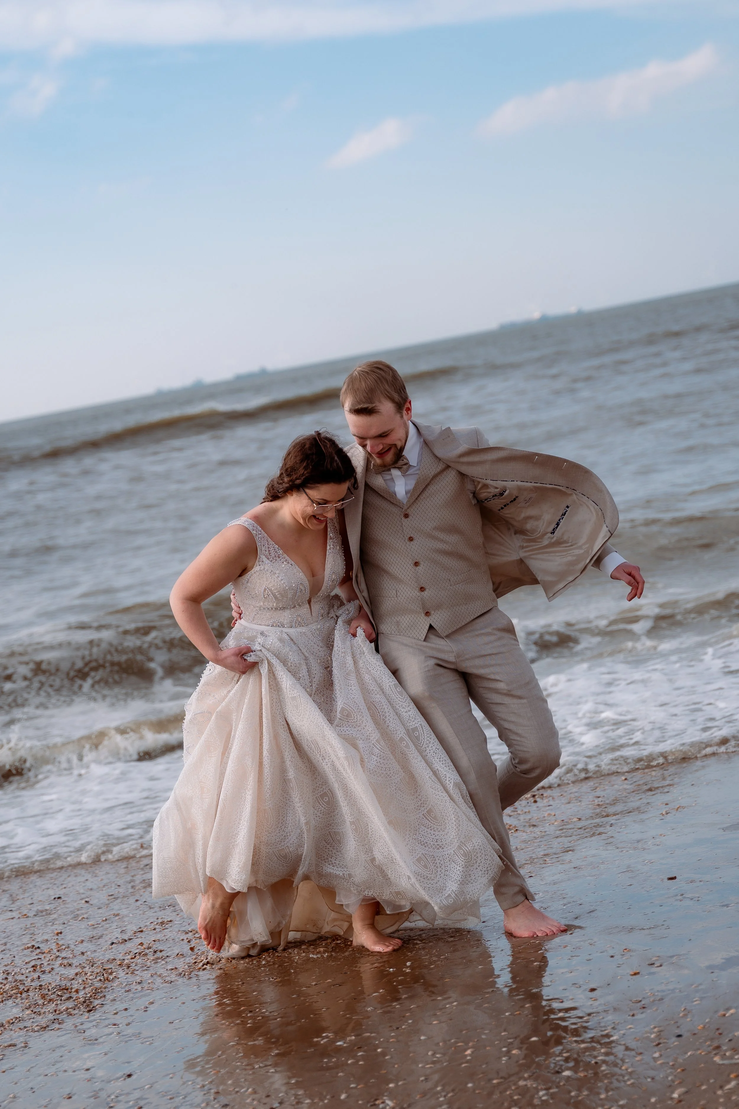 A couple in wedding attire barefoot on the beach, smiling and walking through shallow water, enjoying a moment together.