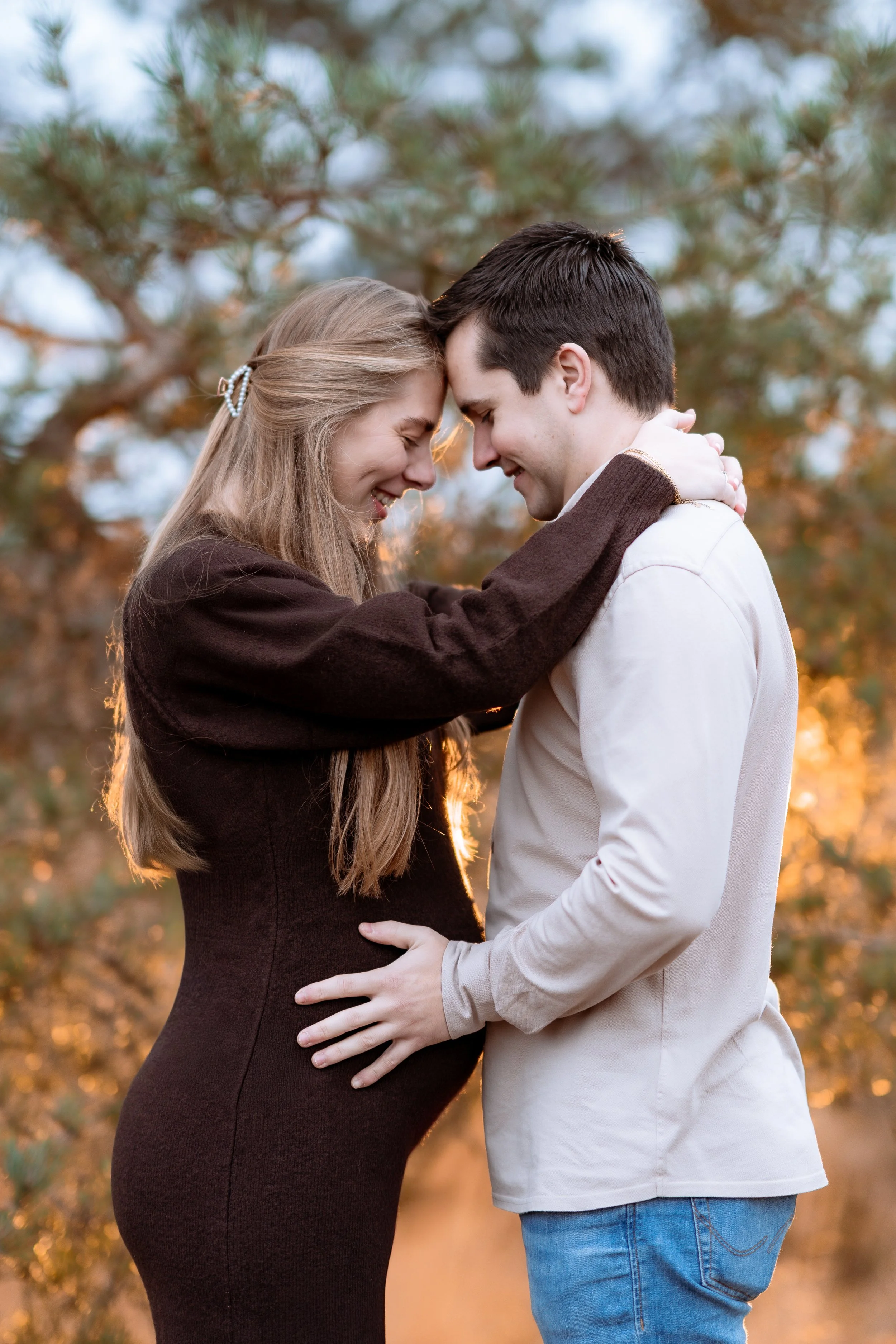 A couple stands outdoors with their foreheads touching, smiling, and embracing. The woman appears to be pregnant, and they are surrounded by trees with a sunset glow in the background.