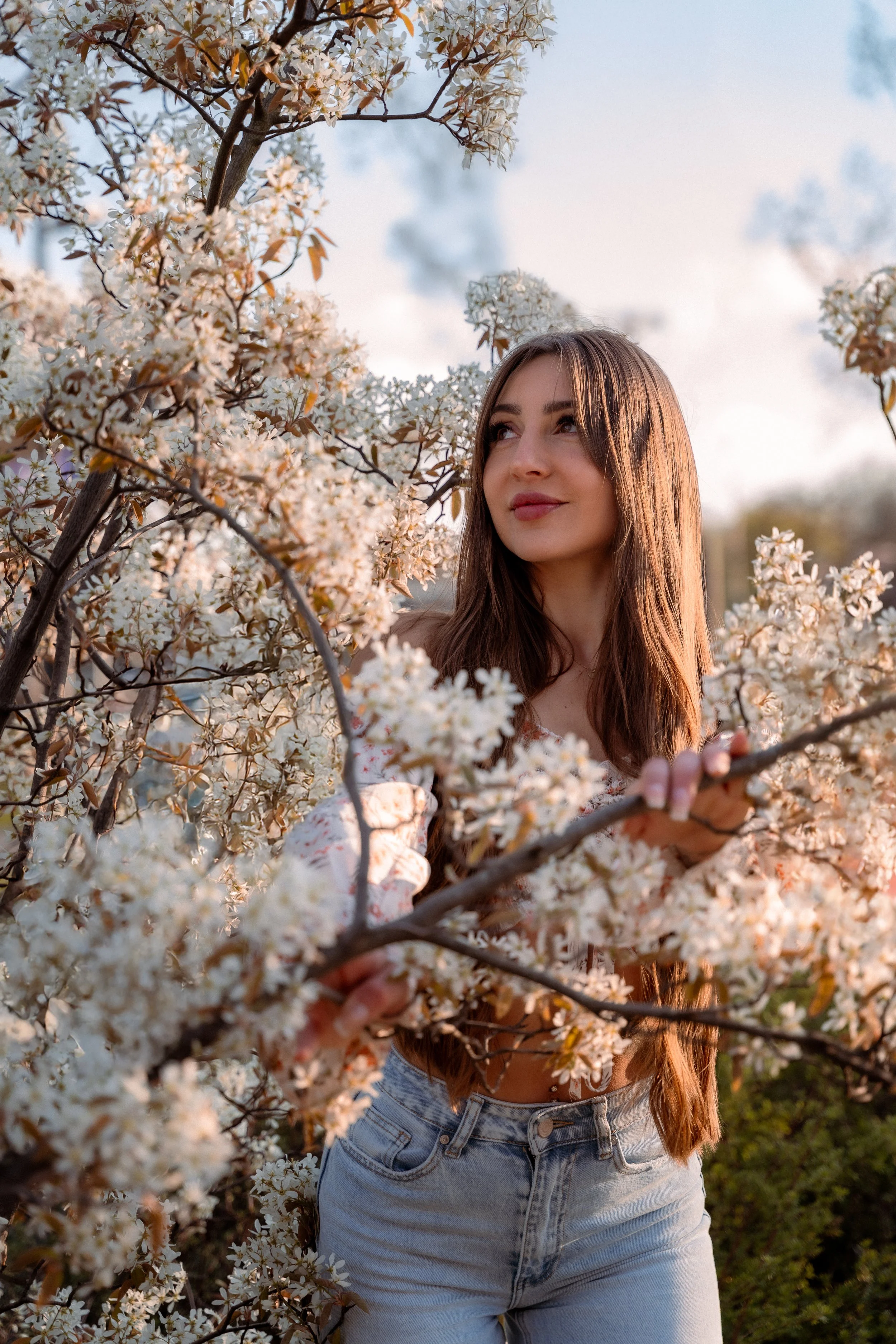A young woman with long brown hair standing among white blossoming flowers on a tree, looking to the side with a gentle expression.