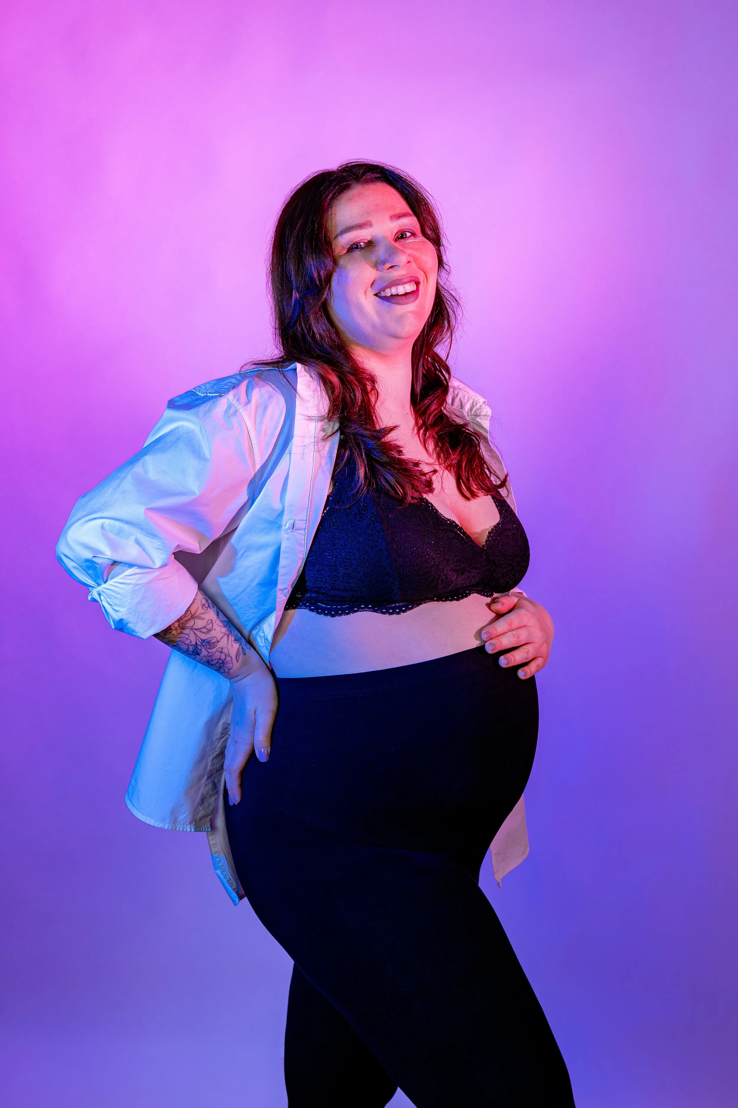A woman with long brown hair wearing a black bralette and black pants, smiling with her hand on her stomach, standing against a purple gradient background.
