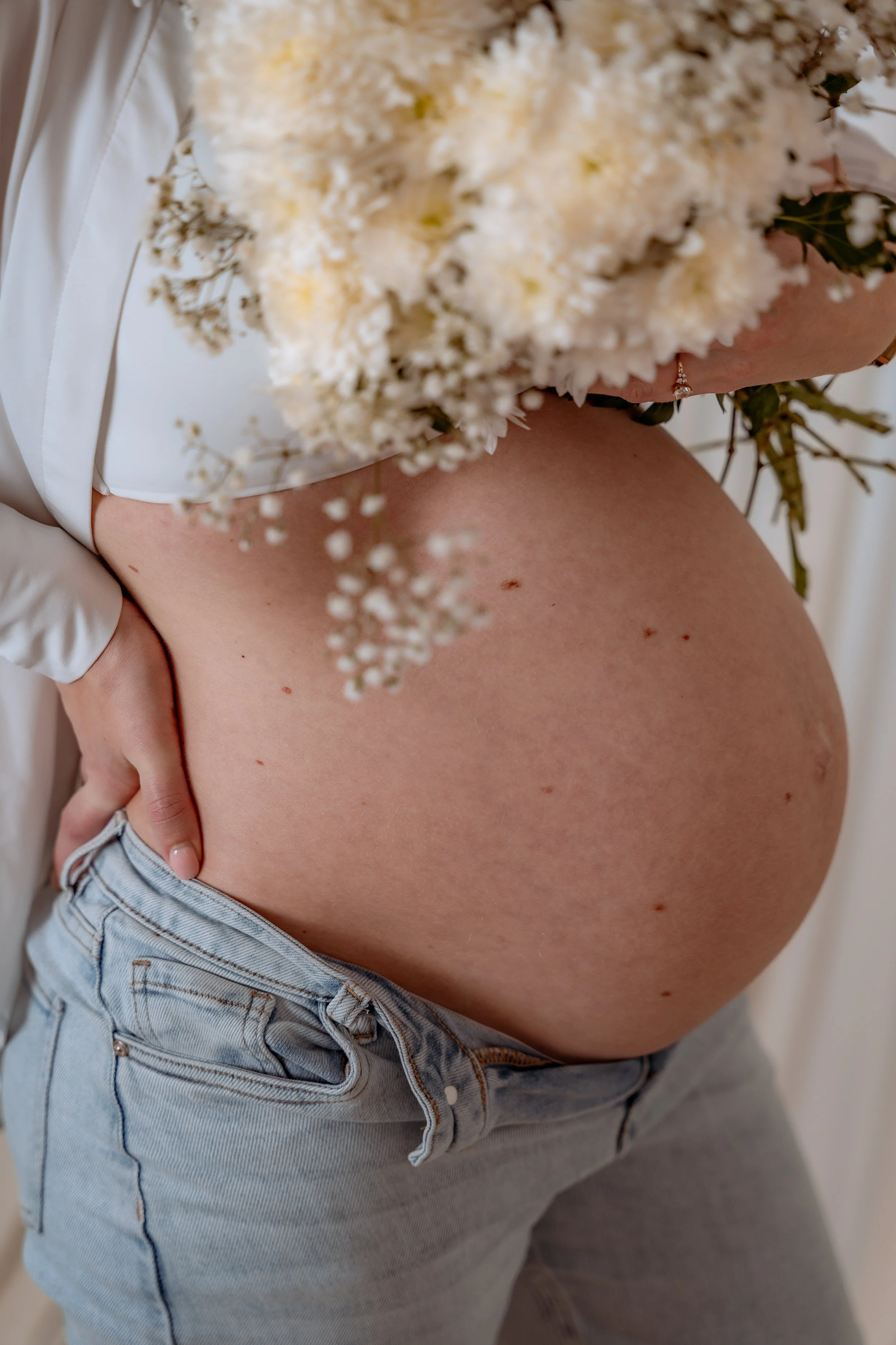 Close-up of a pregnant woman holding a bouquet of white flowers, wearing a white top and light wash jeans.