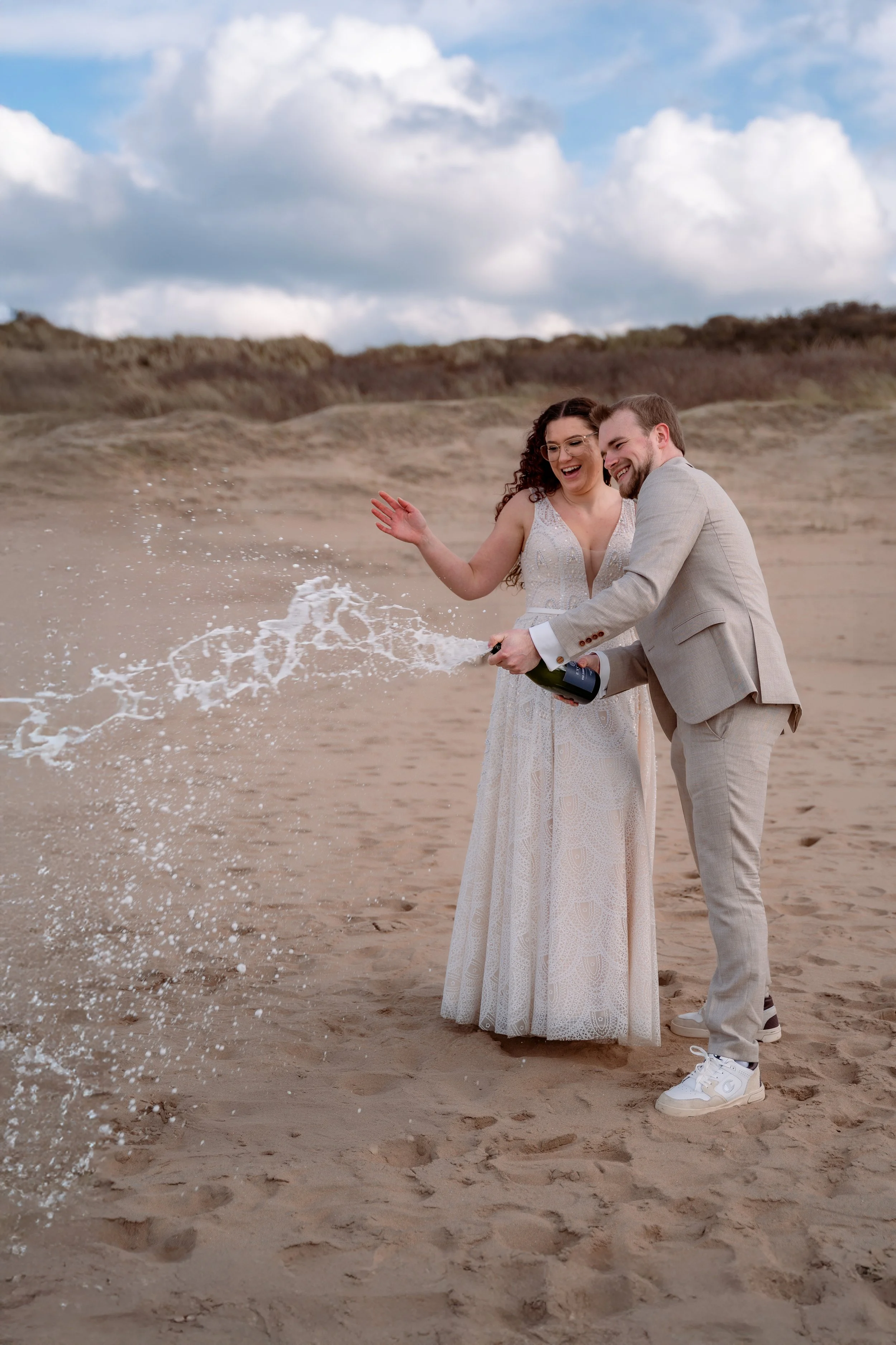 A couple dressed in wedding attire celebrating on a beach as they open a bottle of champagne, with the champagne spraying into the air.