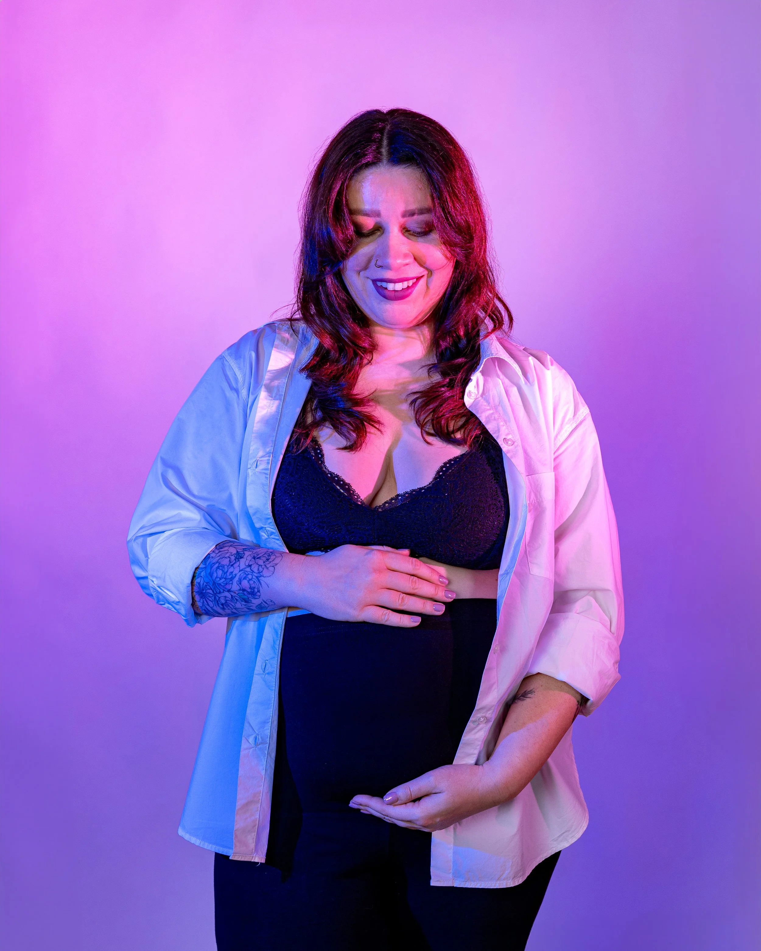 A smiling woman with dark hair, tattoos on her arms, wearing a black lace top and light-colored shirt, standing against a gradient purple background.