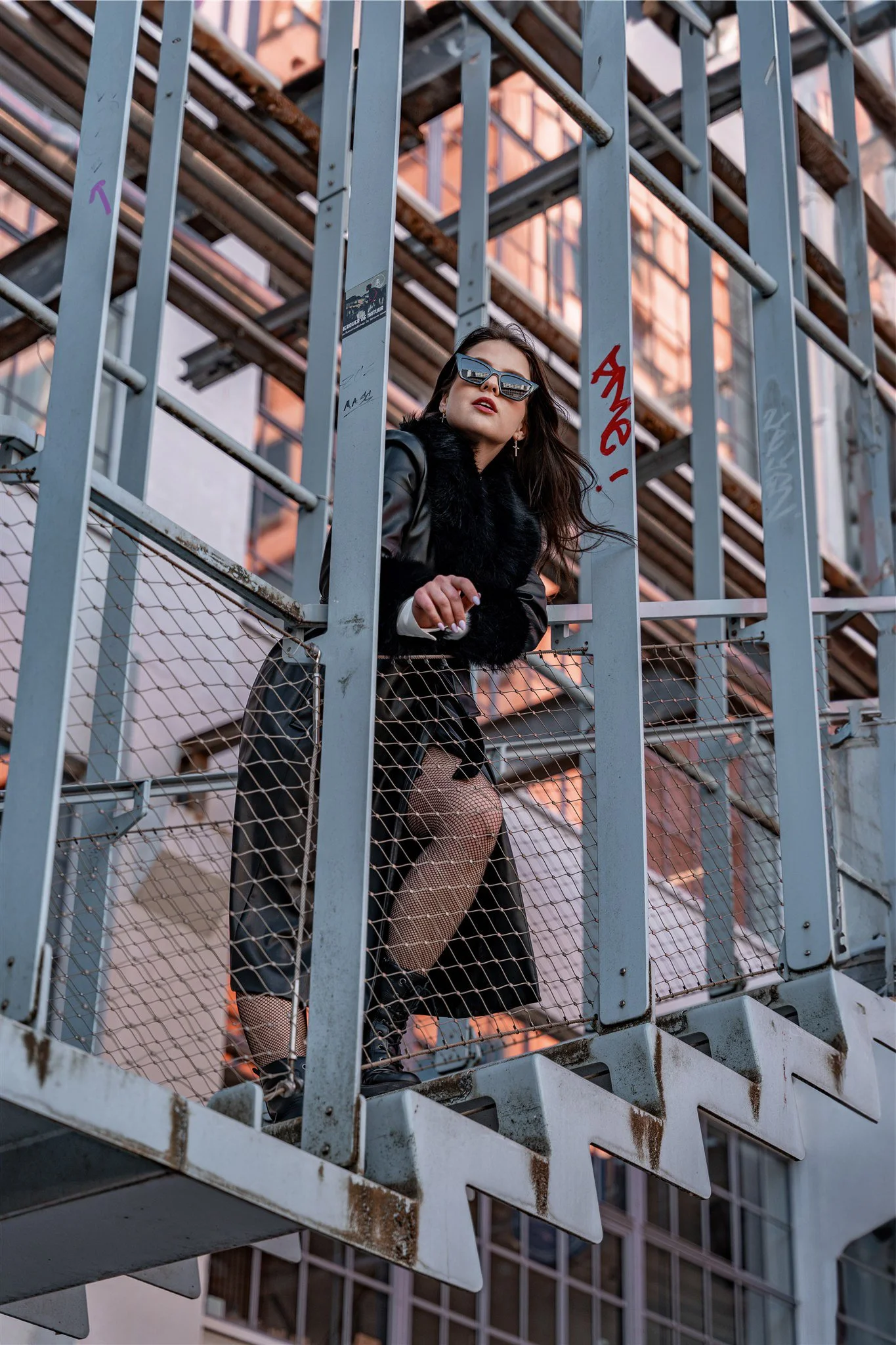 A woman in black clothing and fishnet stockings stands on a fire escape with a city skyline in the background. She wears sunglasses and poses confidently.