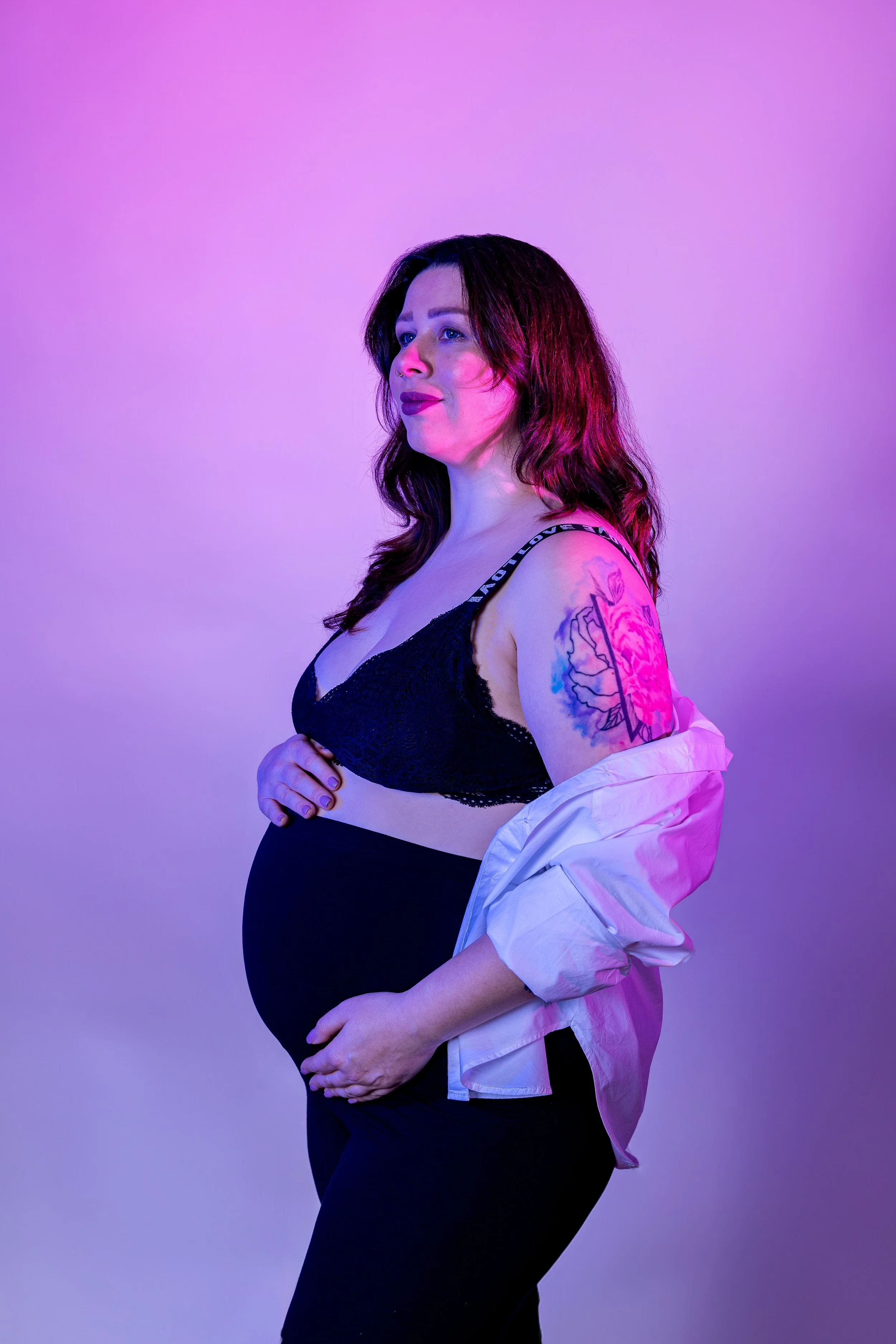 A pregnant woman with long brown hair, wearing black lace lingerie and a white unbuttoned shirt, posed in front of a pink and blue gradient background, with a tattoo on her upper arm.