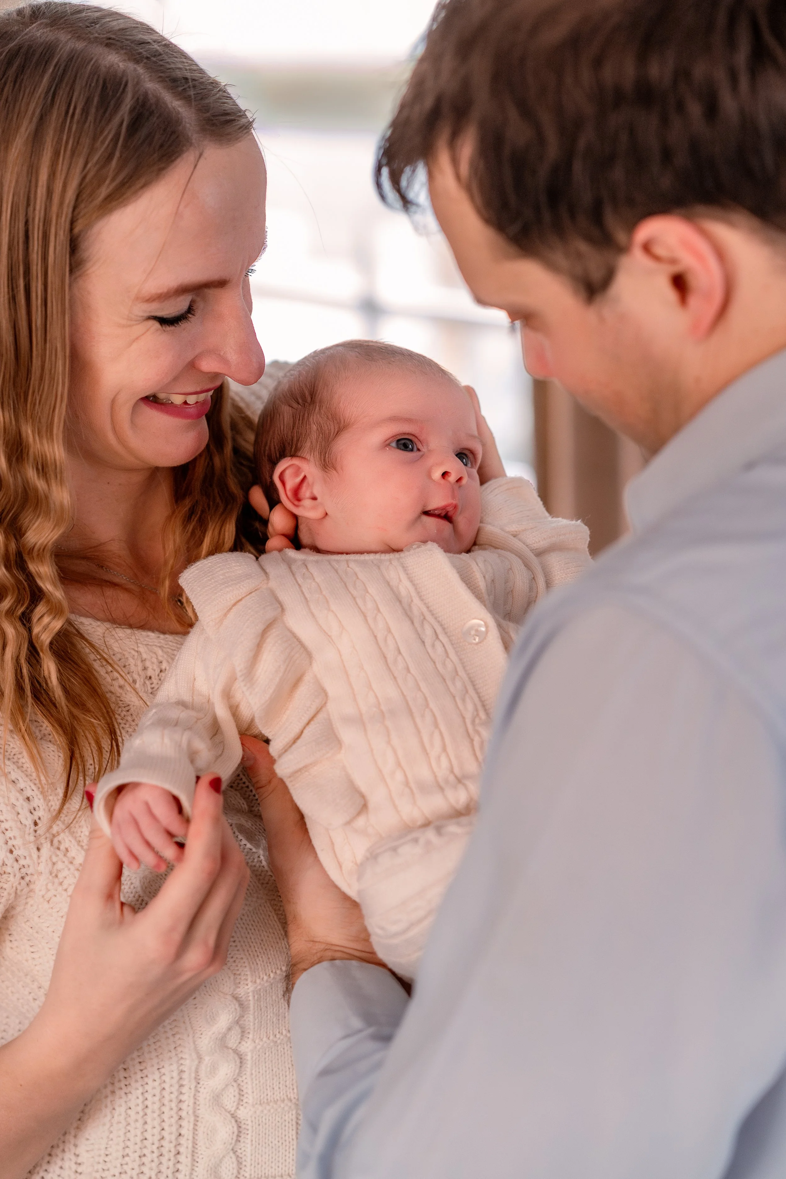 A happy family with a mother, father, and their baby girl at home. The mother is holding the baby and smiling. The father is looking at the baby, who is lying on her back and staring at her father.